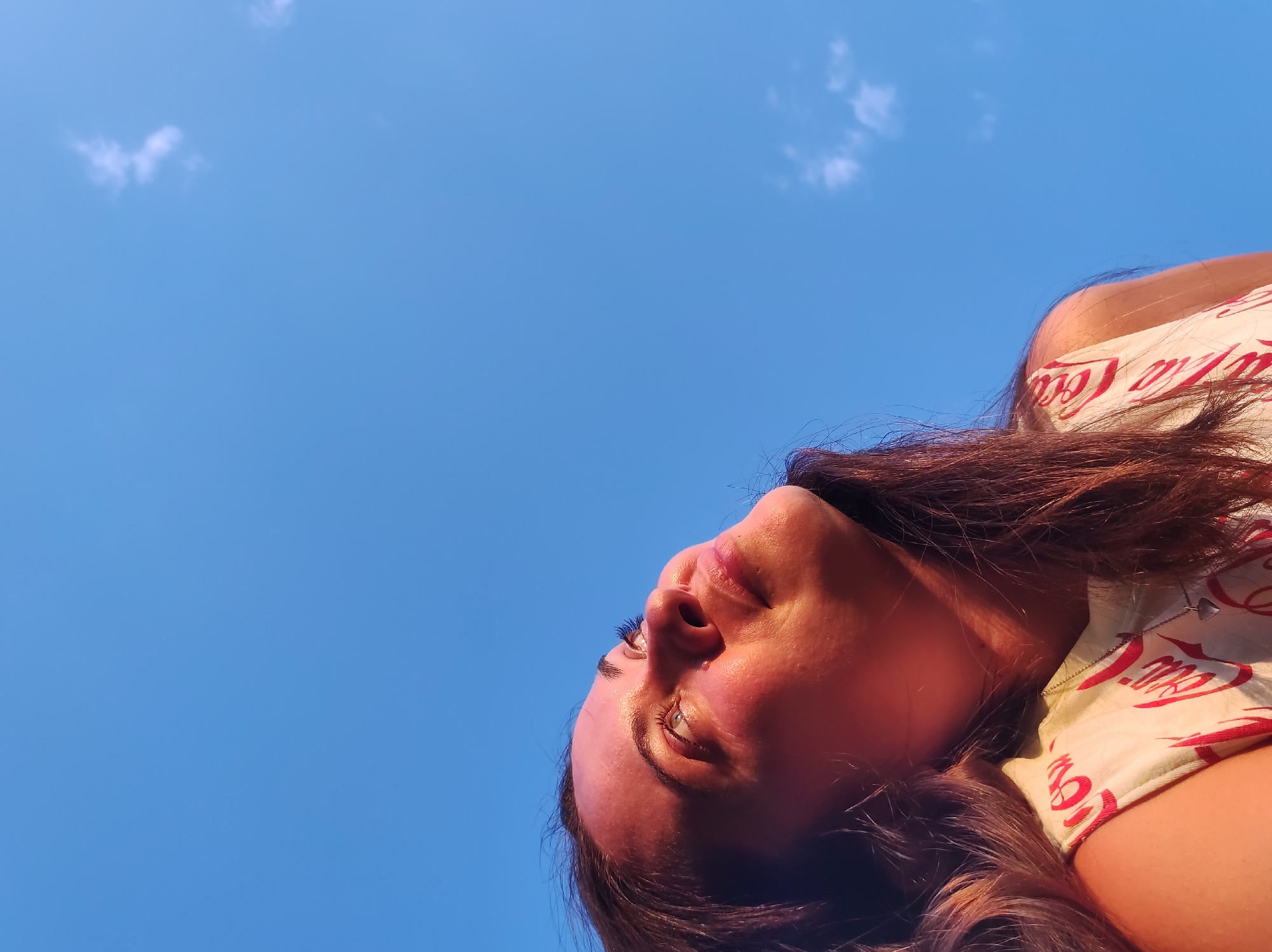 A close-up, low-angle shot features a young woman's face and upper body against a bright blue sky with sparse white clouds. The woman's head is tilted upwards and to the left, with her eyes looking upwards. Her hair is dark brown and cascades around her face and shoulders. She is wearing a light-colored shirt with a repeated red logo, which appears to be "Coca-Cola". The lighting suggests it is daytime, possibly late afternoon, with sunlight illuminating the side of her face. The image is cropped tightly, focusing on the woman and the sky. There are no discernible landmarks or elements that would place this image specifically in the city of Фоки, Russia.