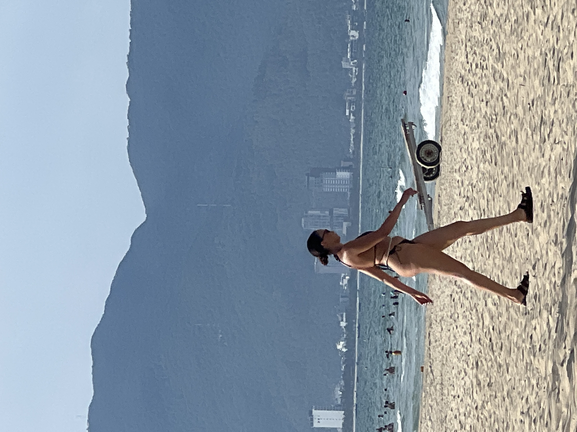 The image captures a side view of a woman in a bikini walking on a sandy beach in Da Nang, Vietnam. She is walking from left to right across the frame, with her right leg extended forward and her left leg bent at the knee. Her hair is pulled back into a bun, and she is wearing sunglasses. Behind her, the ocean is visible with several people swimming and some boats. In the distance, a coastline with buildings and mountains can be seen, characteristic of Da Nang. A small boat trailer with two wheels is positioned on the sand to the woman's right, partially obscured by her. The sky is clear and a pale blue.