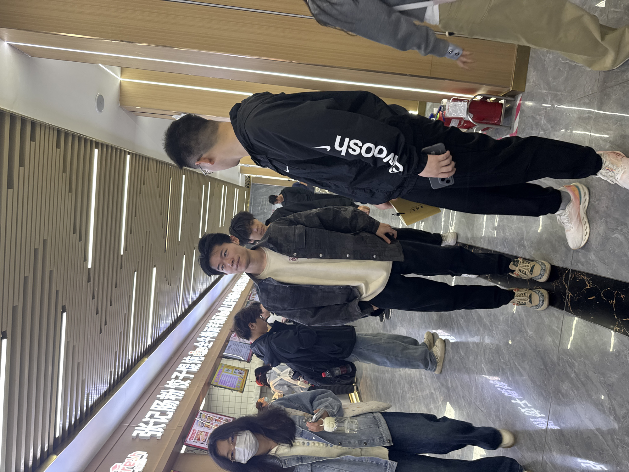 The image shows a line of people, primarily young adults, waiting in a food service establishment. The queue forms along a wall with menu boards featuring Chinese characters, indicating the type of food offered. The location is likely Zhongtun, China, based on the text on the menu.
The people are dressed in casual attire, including denim jackets, hoodies, and t-shirts. Some are wearing masks.
The environment is brightly lit with modern recessed lighting, suggesting an indoor space. The floor is tiled in a dark marble pattern.