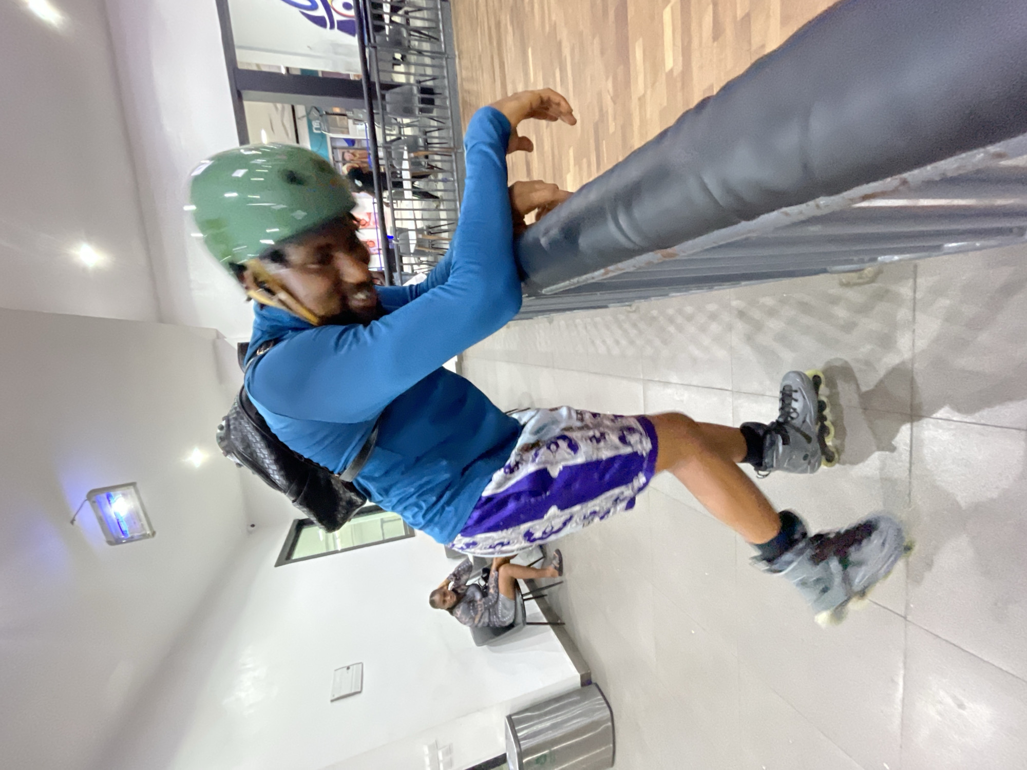 The image depicts a person wearing rollerblades, a helmet, and a backpack, leaning on a railing in what appears to be an indoor roller skating rink or a recreational facility. The person is dressed in a blue long-sleeved shirt and patterned shorts, with their head bent down, possibly taking a break or observing something. In the background, another person is seated on a chair, and there are tables and chairs visible, suggesting a seating or lounge area. The flooring is a light-colored tile, and a metallic trash can is present near the seated person. The lighting appears to be from overhead fixtures. The overall environment suggests a public space designed for recreational activities. The location is specified as Moba, Nigeria.