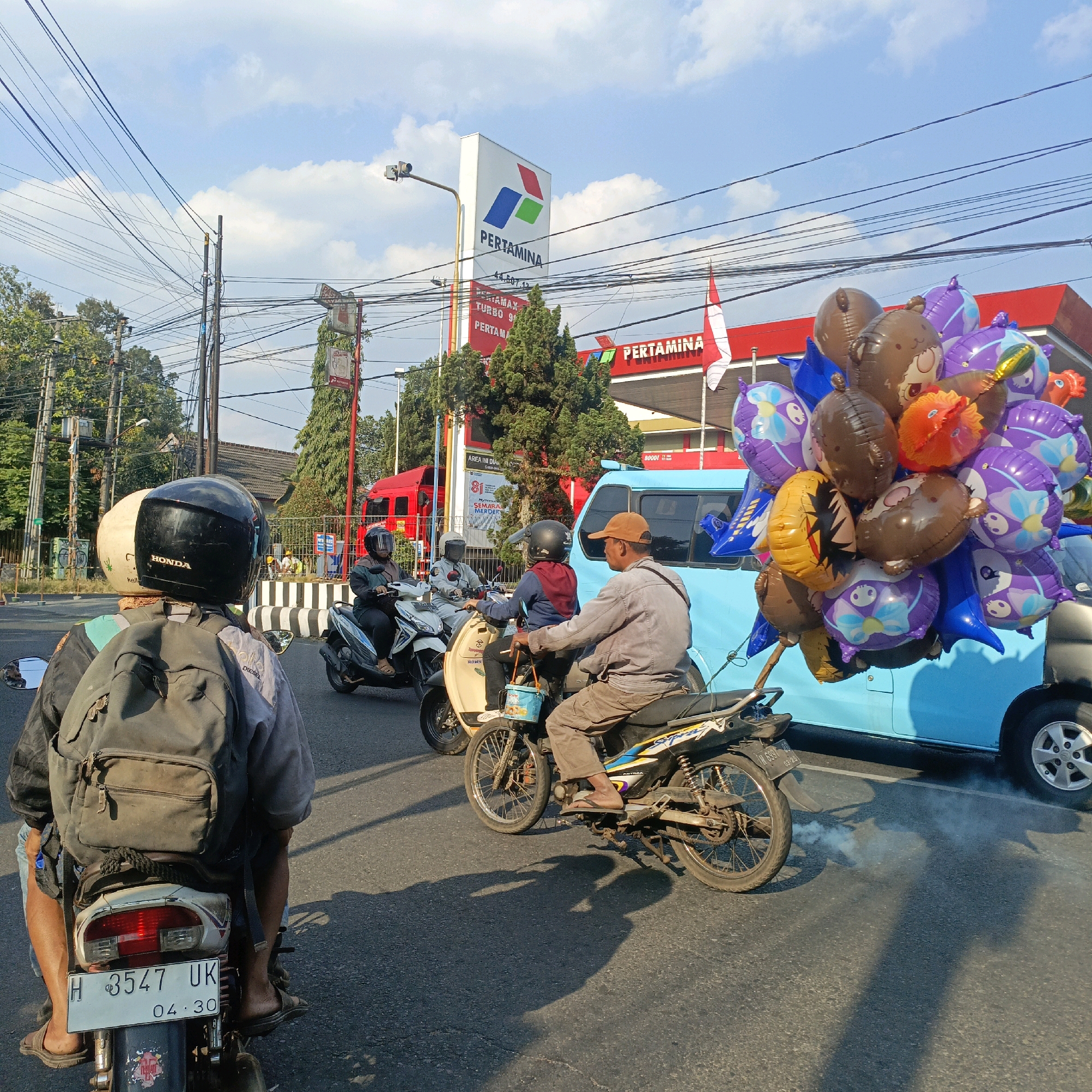 The image depicts a street scene in Salatiga, Indonesia, likely near a Pertamina gas station. In the foreground, two people are riding a motorcycle. The person in the back is wearing a black helmet and a backpack. The person in front is wearing a brown cap and khaki pants. The motorcycle is emitting a cloud of smoke. To the right, a light blue vehicle is parked, adorned with a large cluster of colorful balloons, some featuring cartoon characters like bears and flowers, and others being solid colors like brown, orange, and purple. Behind the vehicle and balloons, the bright red building of a Pertamina gas station is visible, with its prominent signage. Several other motorcycles and riders are present in the mid-ground, navigating the street. The sky above is a clear blue with scattered white clouds, and a network of overhead electrical wires crisscrosses the scene. The overall impression is a busy urban street with everyday activity, including commerce represented by the balloons and the gas station.