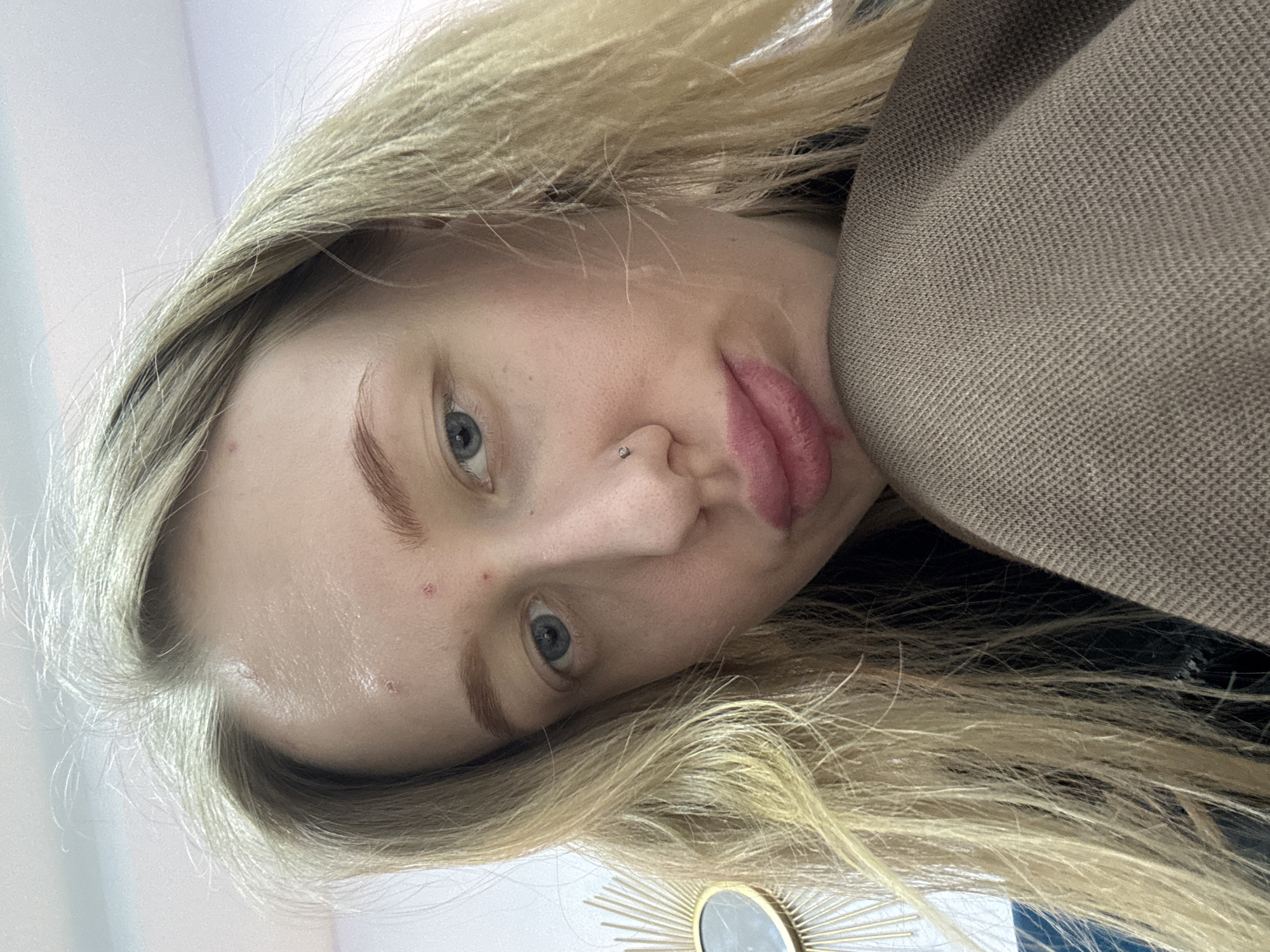 A close-up, slightly rotated, selfie-style image shows a young, fair-skinned person with blonde hair and blue eyes. The person has a nose piercing, slightly parted red lips, and visible skin blemishes. A brown textured garment is visible on the right side of the frame. In the bottom of the picture there is a decorative mirror. There is no information given in the picture to indicate a city or region.