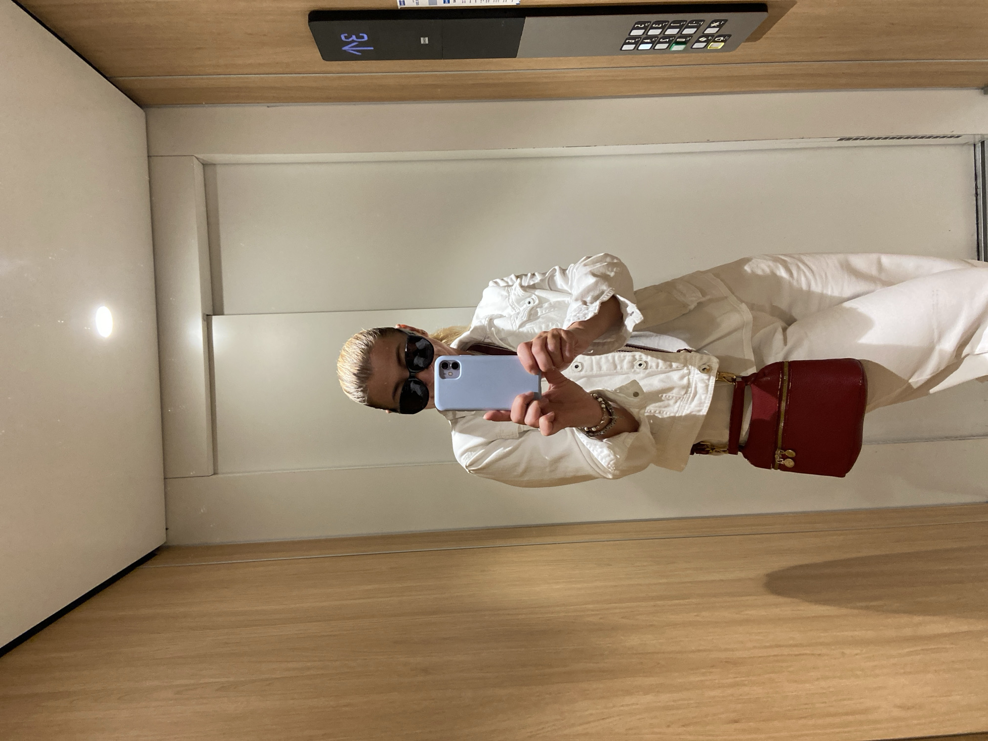 The image is taken in an elevator. The elevator has wood paneling with white walls. At the top of the image are elevator buttons indicating it is on the 3rd floor. The person in the image is facing the mirror and taking a photo of themself. They are wearing white clothing and sunglasses. The image does not provide sufficient information to determine the exact location in Budva, Montenegro.