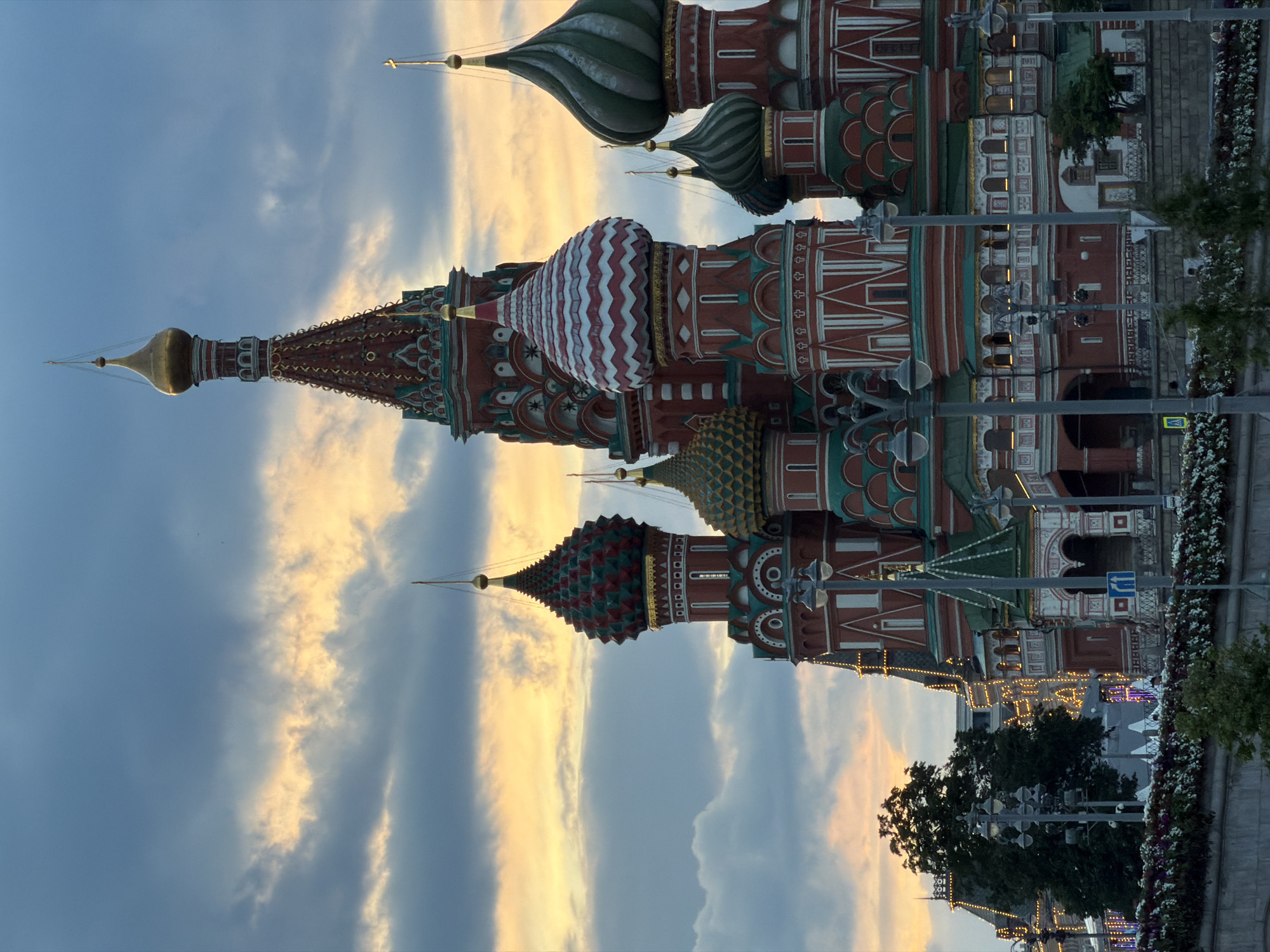 The image displays a captivating view of St. Basil's Cathedral in Moscow, Russia, captured during what appears to be either late afternoon or early evening. The iconic cathedral stands prominently, showcasing its vibrant and intricate architecture against a partly cloudy sky.
The cathedral features a collection of distinctive onion domes, each adorned with unique patterns and colors. The central dome is topped with a golden spire, while others exhibit diverse designs, including a prominent white and red striped pattern, and scales of green and red. The main structure of the cathedral is characterized by reddish-brown walls with ornate white and teal green decorative elements.
The sky is a blend of soft blue and scattered clouds, with warm, golden-orange light filtering through, particularly on the cloud edges, suggesting the sun is low on the horizon. This lighting enhances the cathedral's rich colors and textures. In the foreground, several streetlights with decorative lamps are visible, along with green trees and what appear to be flowerbeds or planted areas, indicating a public space like Red Square. No individuals are clearly visible in the frame. A small yellow pedestrian crossing sign and a blue directional sign are faintly discernible in the lower right, but any text on them is illegible. The overall scene conveys a sense of architectural grandeur and historical significance under a picturesque sky.