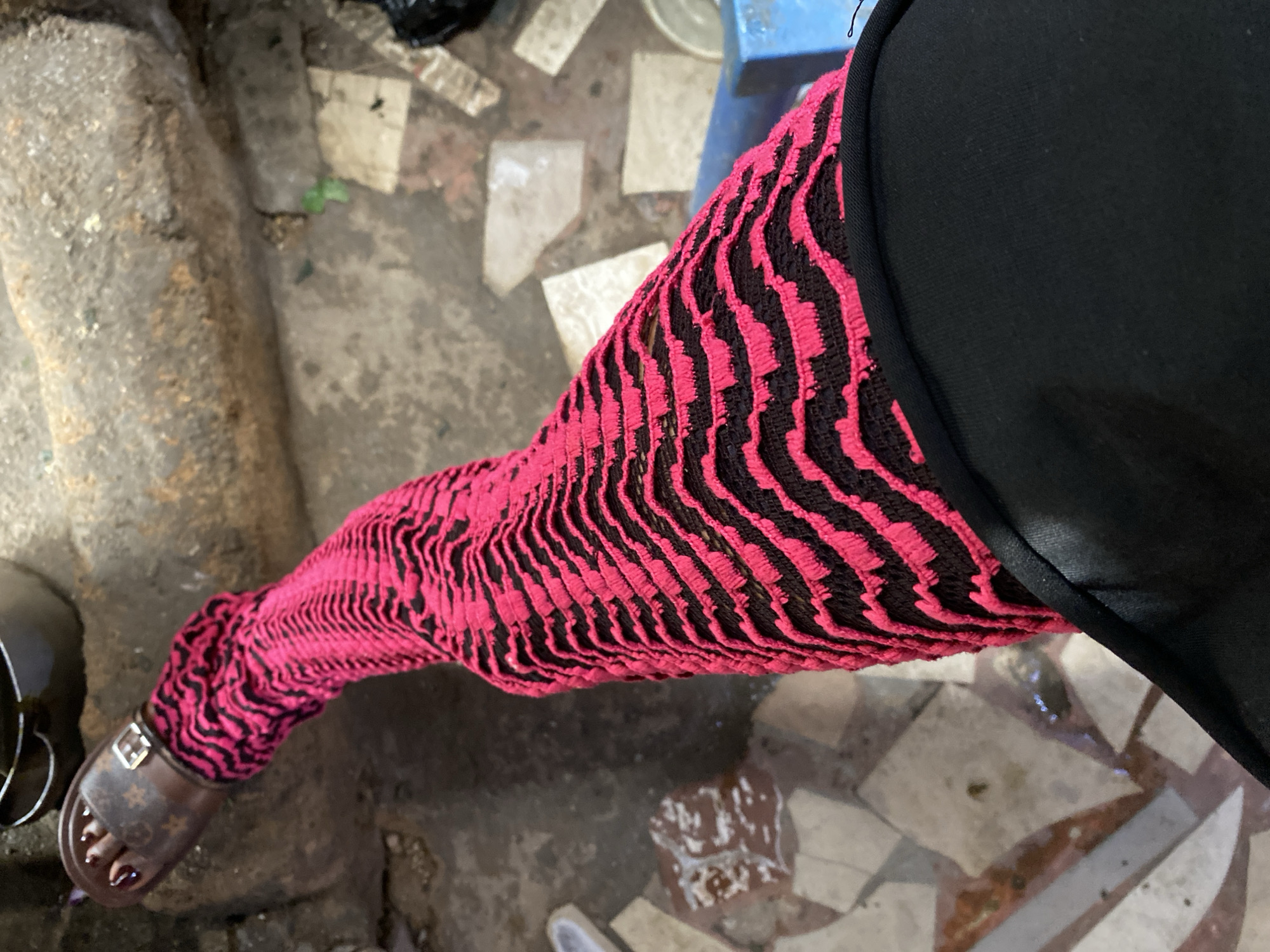 The image shows a close-up of a person's leg wearing a patterned stocking. The stocking is predominantly pink with black zig-zag patterns. The person is also wearing a brown sandal with a gold buckle, featuring a monogram pattern, on their right foot. The background appears to be an outdoor environment, possibly a street or courtyard, with a rough stone surface, broken tiles, and some debris. A blue object is partially visible in the lower right corner. The overall impression is one of a candid snapshot of fashion in an urban setting. The location in Jos, Nigeria, suggests a context of everyday life in that West African city, hinting at the cultural and stylistic influences present there.