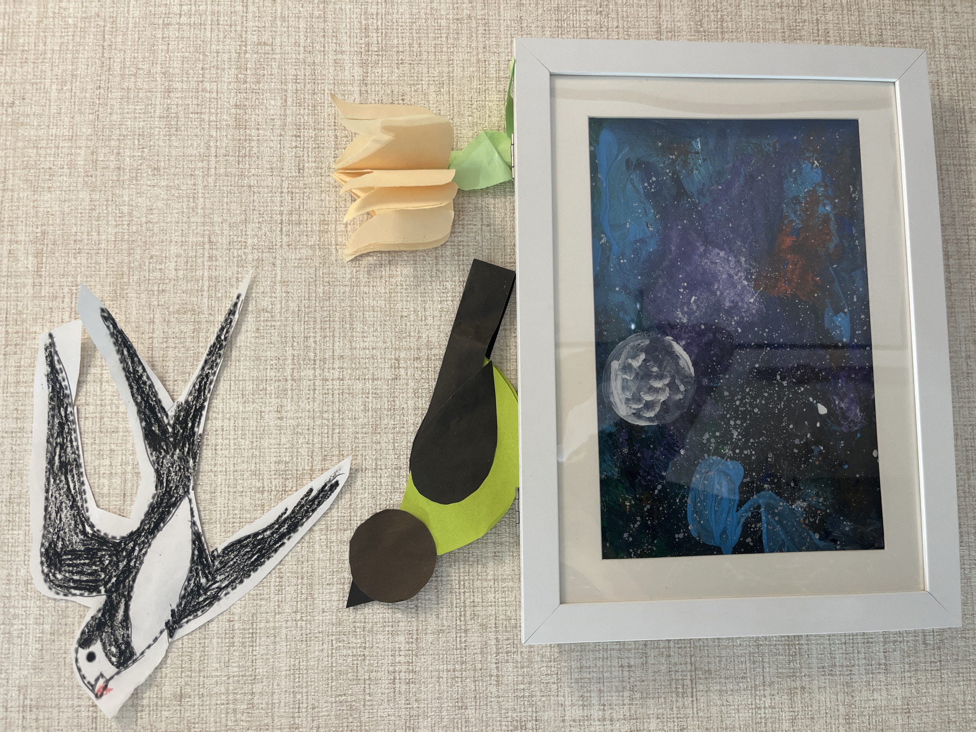 The image displays a wall decorated with various art pieces, including a drawing of a swallow, a paper cutout bird, a paper tulip, and a framed painting. The swallow drawing, rendered in black and white crayon, is in flight with its wings spread. Below it, a brown and green paper bird cutout is perched. To the right of the bird cutout is a peach-colored paper tulip with a green stem. A white-framed painting hangs below these elements, depicting a night sky with a large moon and colorful, abstract shapes that could represent nebulae or galaxies. The wall itself has a textured, light-colored surface. There are no people visible in the image. The scene appears to be an indoor setting, possibly a child's room or a decorative display. There is no indication of the location being Noyabrsk, Russia.