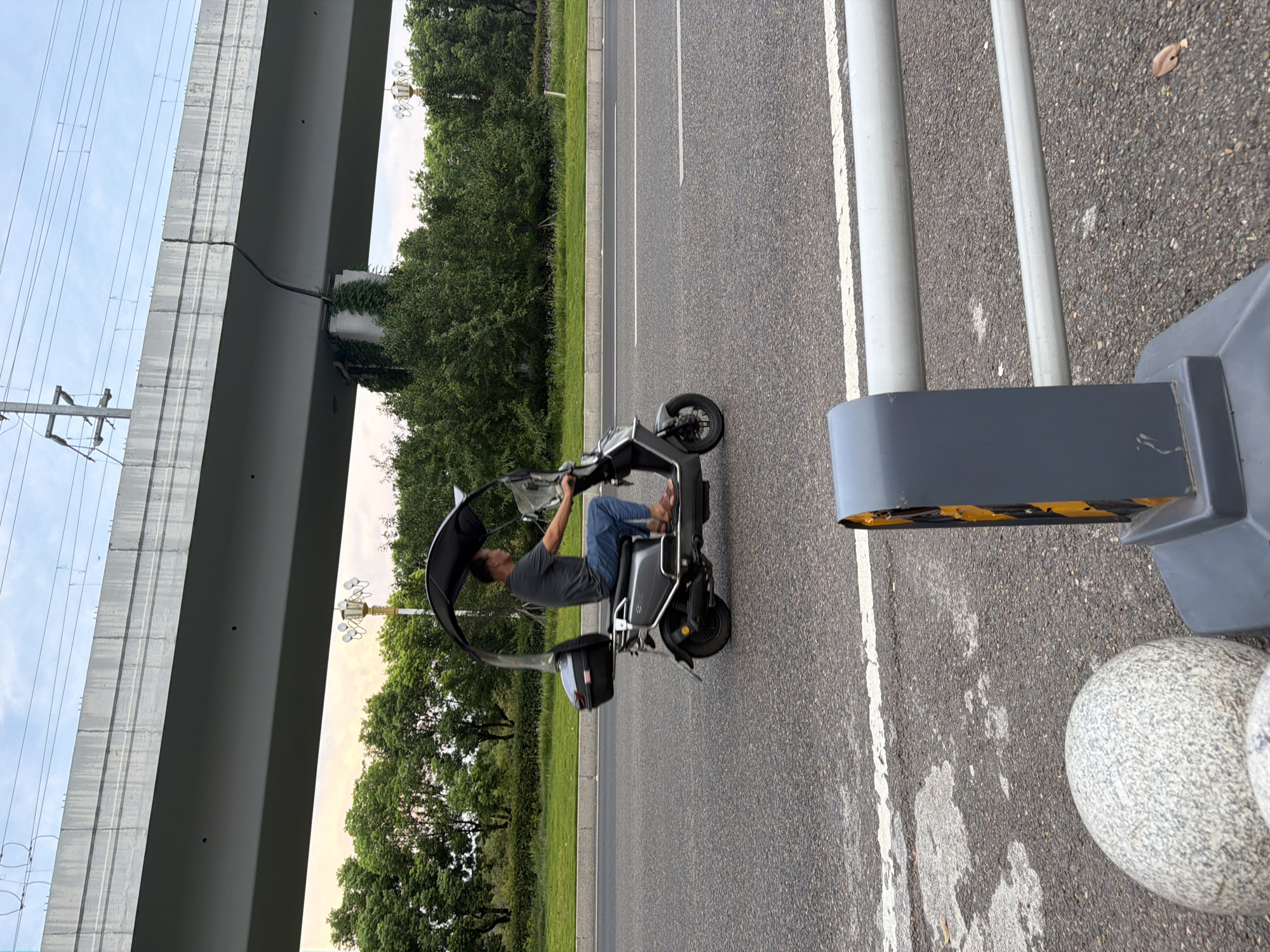 A middle-aged East Asian male is riding a dark gray electric scooter on a paved road. He is wearing a dark gray t-shirt and blue jeans, and brown sandals. The scooter has a black canopy extending over the rider and a black storage box mounted behind the seat. The scooter is in the right lane of a two-lane road. In the background, there are trees and a large concrete overpass. The sky is overcast. In the foreground, there is a gray barrier with yellow reflective strips and a round gray stone. The image appears to have been taken in the city of Jiangjin, China.