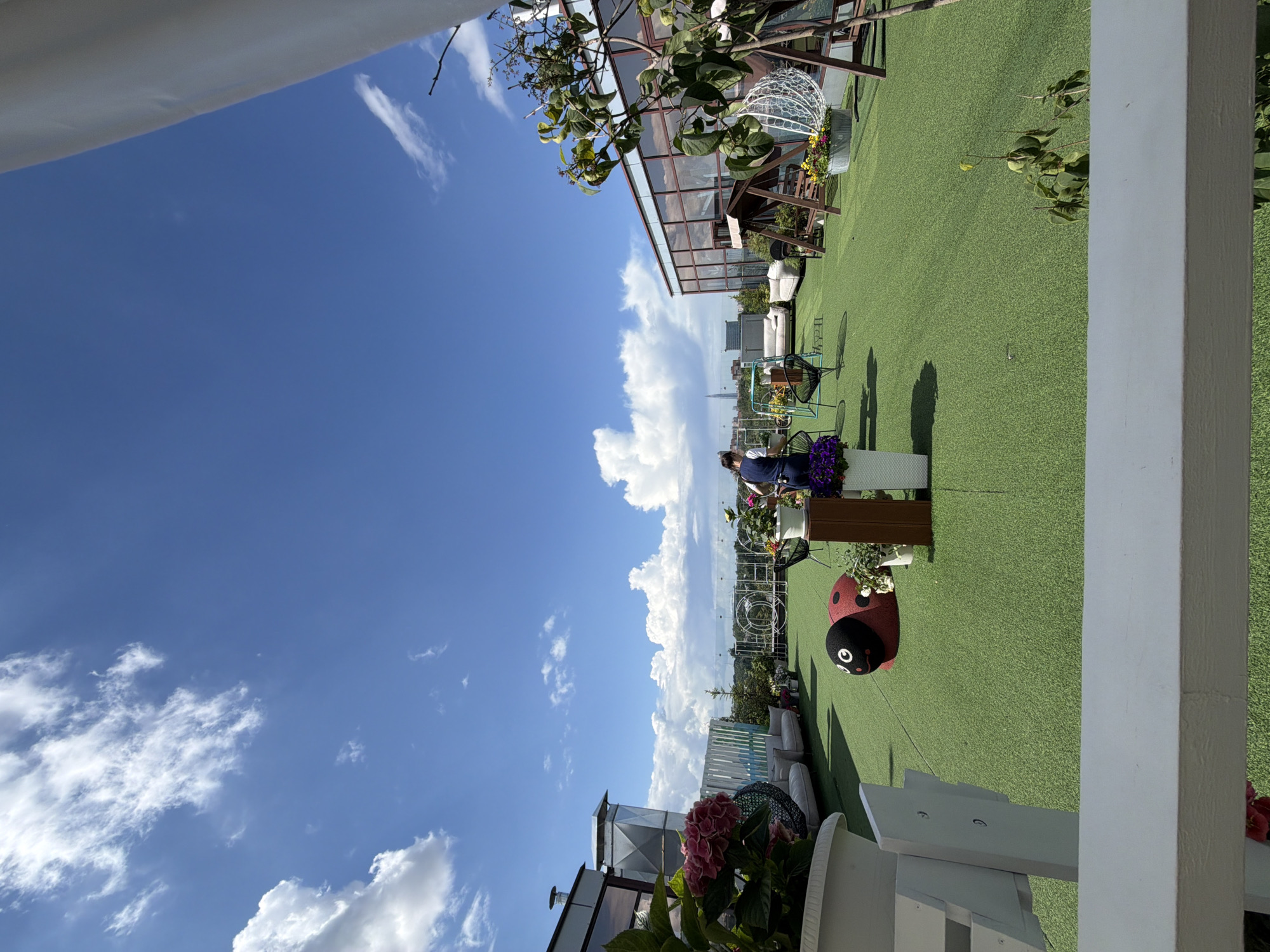 This is a wide-angle, eye-level shot of a rooftop terrace in Saint Petersburg, Russia, on a sunny day with scattered clouds. The terrace is covered with artificial green turf and features various seating areas and decorative elements.
In the foreground, a white wooden railing and planter are visible. To the left, a large planter contains pink hydrangeas and a black wire decorative sphere. Further back, a red and black ladybug-shaped ottoman is placed on the turf. A person wearing a dark blue uniform is standing near a table with flowers and appears to be arranging them.
The background reveals a cityscape, with several buildings, including a modern glass-fronted structure to the right. A swing set with a wooden frame and a white hanging sphere are also present on the terrace. White sofas and chairs are scattered around the area, suggesting a relaxation or event space. In the distance, a tall, slender skyscraper is visible. The overall impression is of a well-appointed outdoor space with a view of the city.