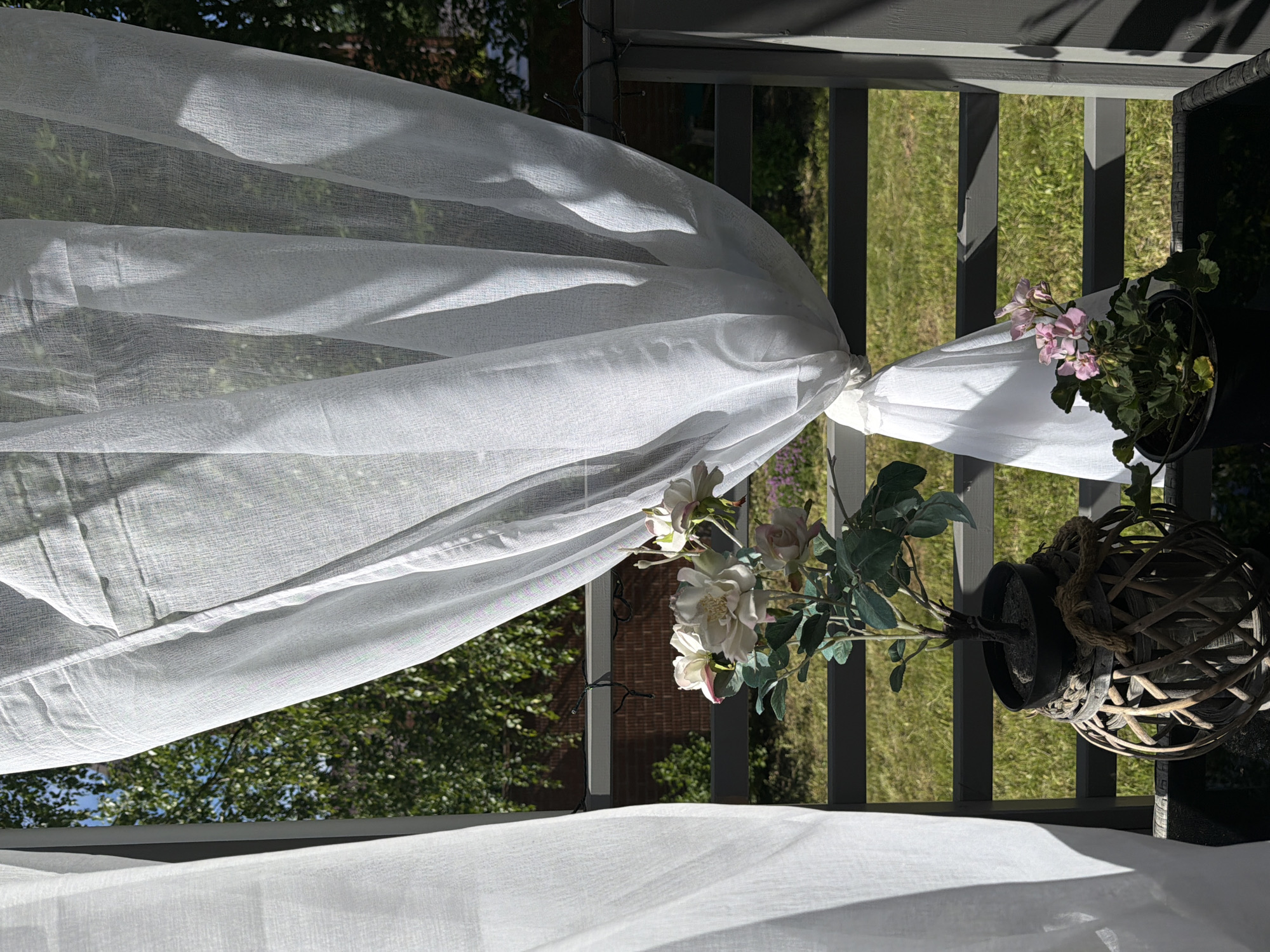The image depicts a close-up view of a white sheer curtain blowing gently in the wind on a balcony. Through the translucent fabric, a glimpse of green foliage and a brown brick wall is visible, suggesting an outdoor setting. In the foreground, two potted plants are arranged on the balcony. The plant on the left features delicate white and pink artificial roses with green leaves, housed in a dark pot that is placed inside a woven wicker basket. The plant on the right displays small pink flowers and green foliage, also in a dark pot. A string of dark-colored fairy lights is draped across the grey railing of the balcony, partially obscured by the curtain. The overall scene suggests a bright, sunny day, possibly in a residential area of Алешкино Дп, Russia, with the natural light creating soft shadows on the curtain and railing. There are no people or animals present in the image.