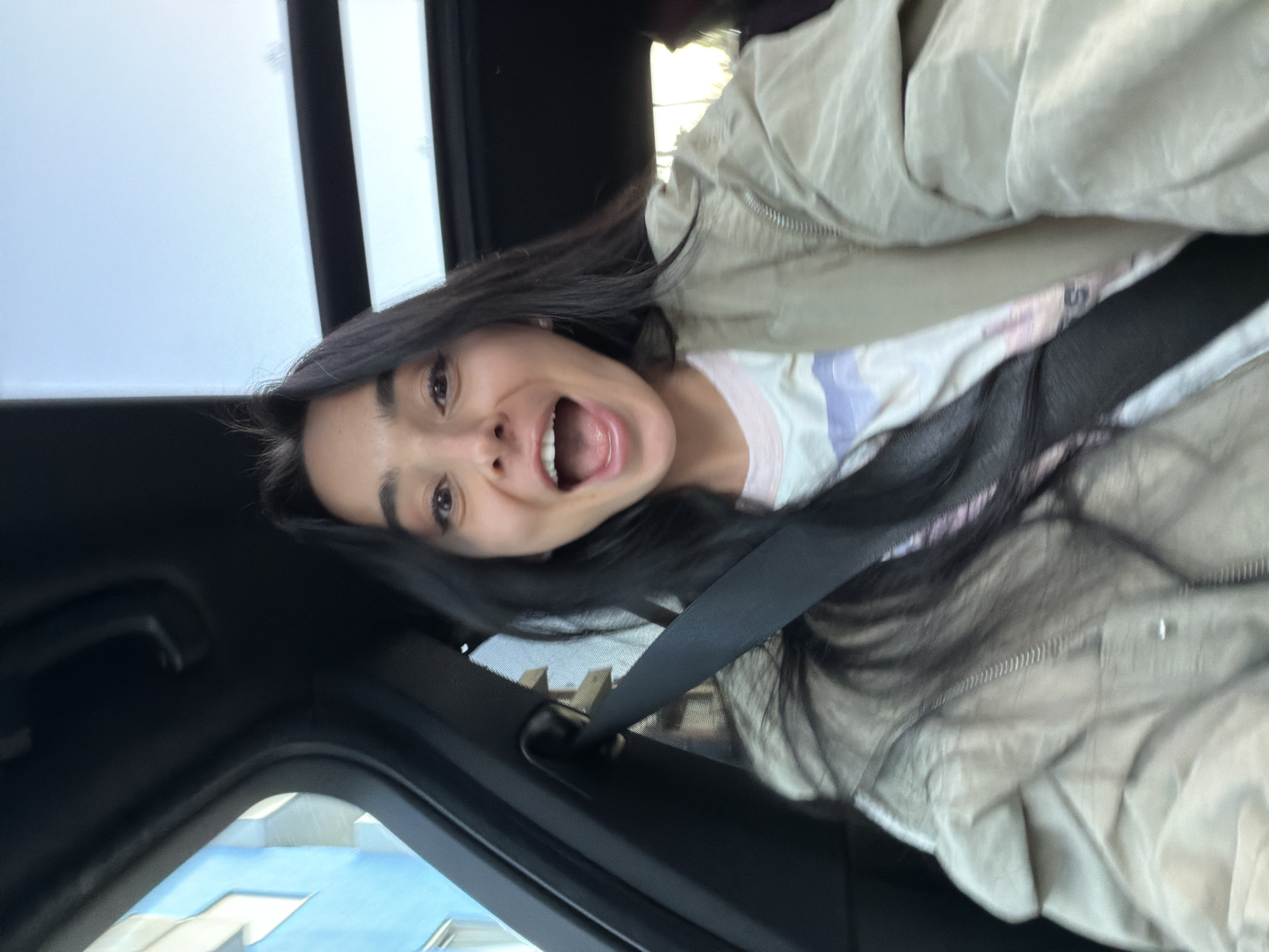 A woman is in the driver's seat of a car, wearing a seatbelt, a light-colored jacket, and a T-shirt with light purple and pink accents. She has long, dark hair and an open mouth. The car's interior is visible, including parts of the dashboard and the sunroof. Outside the car window, a building with blue accents is visible, which may be located in the city of Grad Rijeka, Croatia.