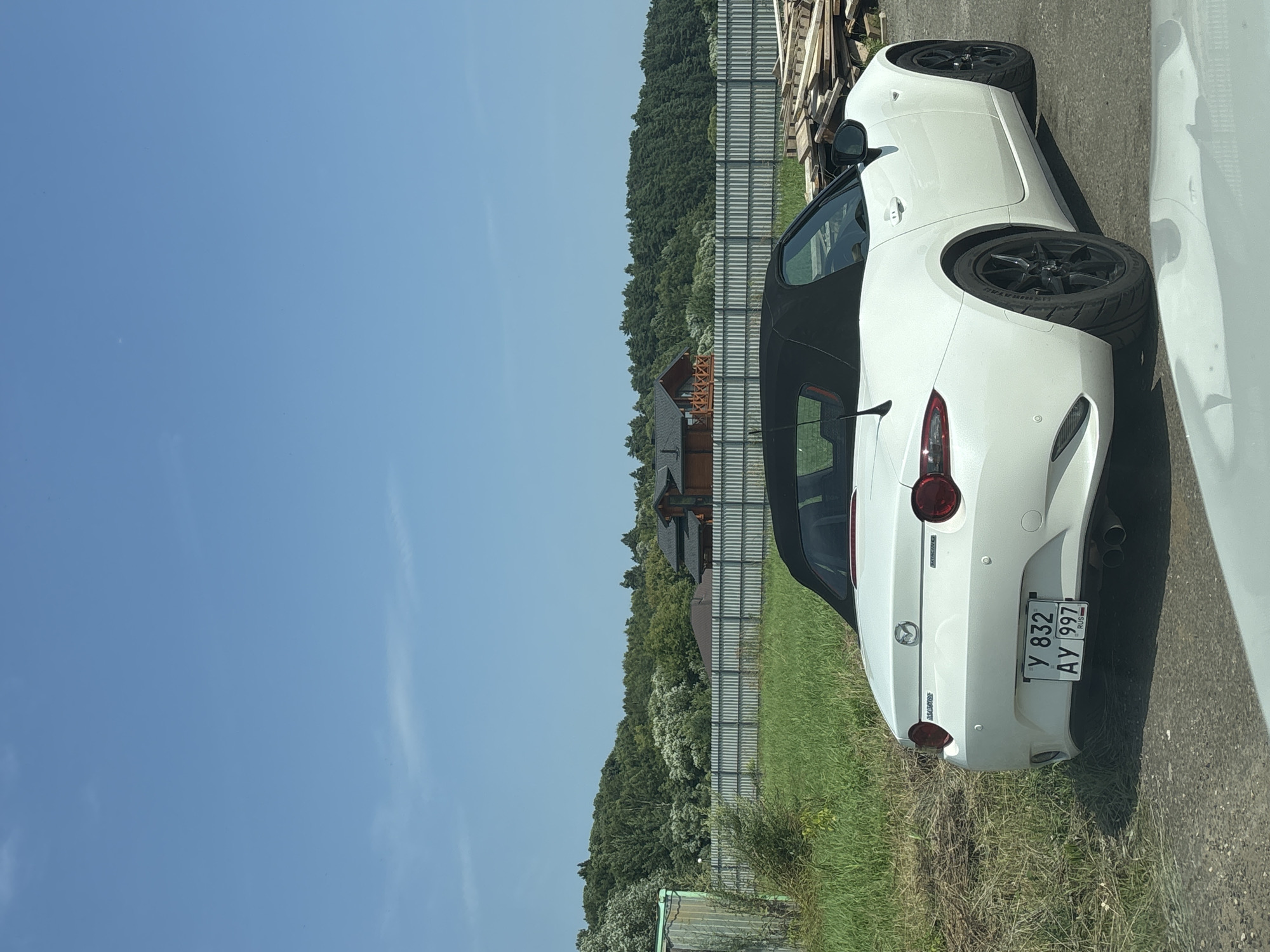 A white Mazda MX-5 convertible is parked outdoors on a sunny day in Zvenigorod, Russia. The car is positioned facing left, with its black soft top up. A license plate with the characters "Y 832 AY 997 RUS" is visible on the rear of the vehicle. In the background, a metal fence separates a grassy area from a wooded landscape with trees and several wooden houses. To the right of the car, a stack of wooden planks is visible. The sky is clear and blue.