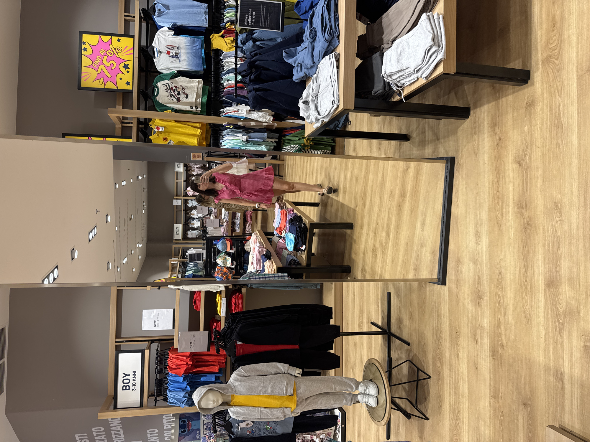 The image depicts the interior of a clothing store, specifically "OVS" in Carini, Italy, as indicated by text in the image. A woman in a pink dress is reflected in a large mirror, taking a photo of herself with her phone. She is standing in an aisle of the store, surrounded by racks of children's clothing and displays. To her left, a mannequin is dressed in a grey tracksuit and white sneakers, positioned on a circular display stand. The store features wooden floors and shelving units. In the background, various clothing items are organized on hangers and shelves, with promotional signs visible. One sign advertises a sale of "FINO AL 75%". Another sign indicates "Nuove collezioni in arrivo" (New collections arriving). The overall scene is well-lit, consistent with a retail environment.