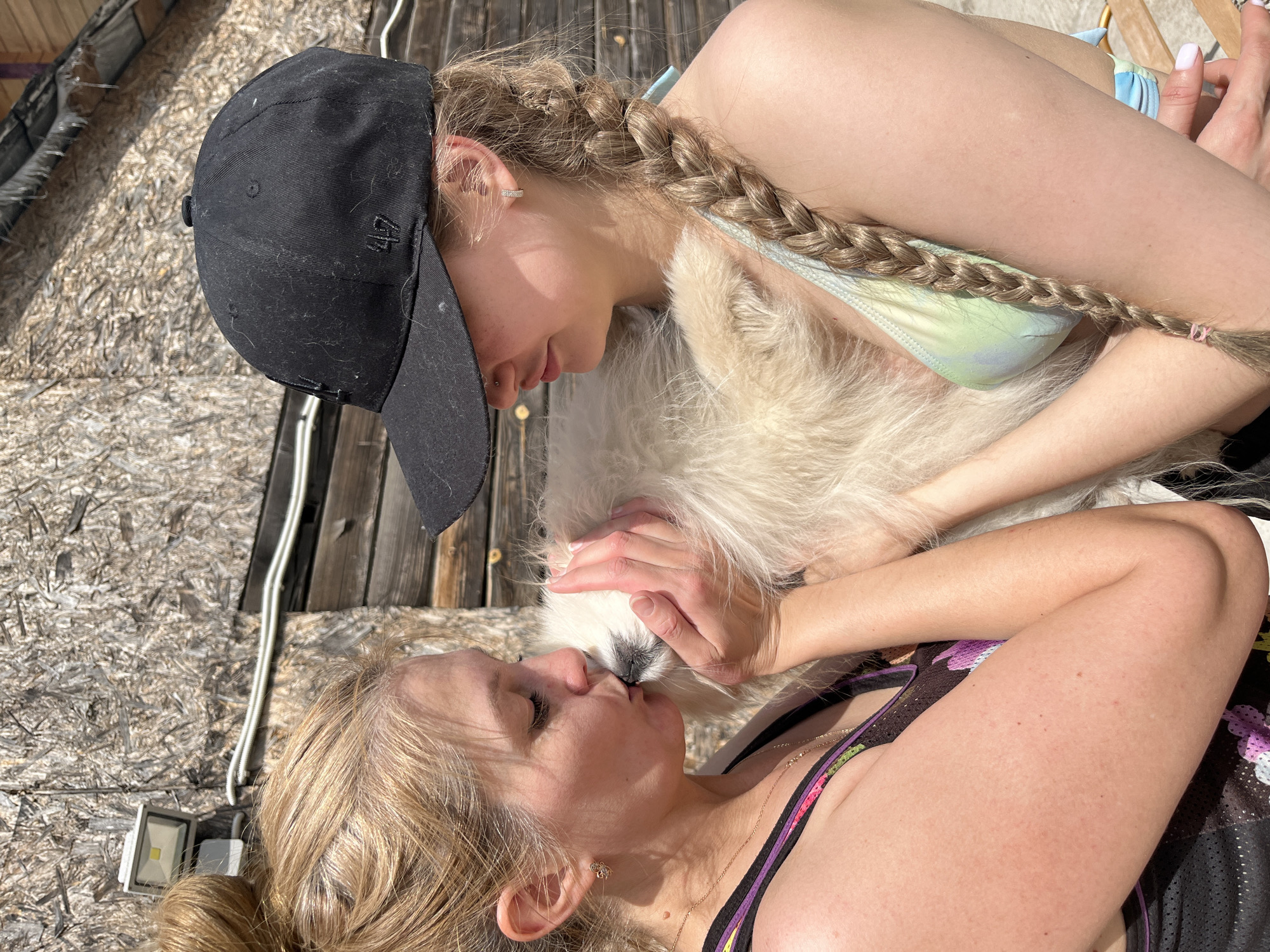 The image shows two women and a white dog outdoors. One woman is lying on her back wearing a dark colored tank top with floral designs and has blonde hair. The second woman is lying over the first, wearing a bikini top and a black baseball cap, with a blonde braid. They are interacting with a fluffy white dog positioned between them, possibly kissing or cuddling the dog. The background appears to be a wooden surface. The location and details provided about Izhevsk, Russia are not discernible from the provided image.