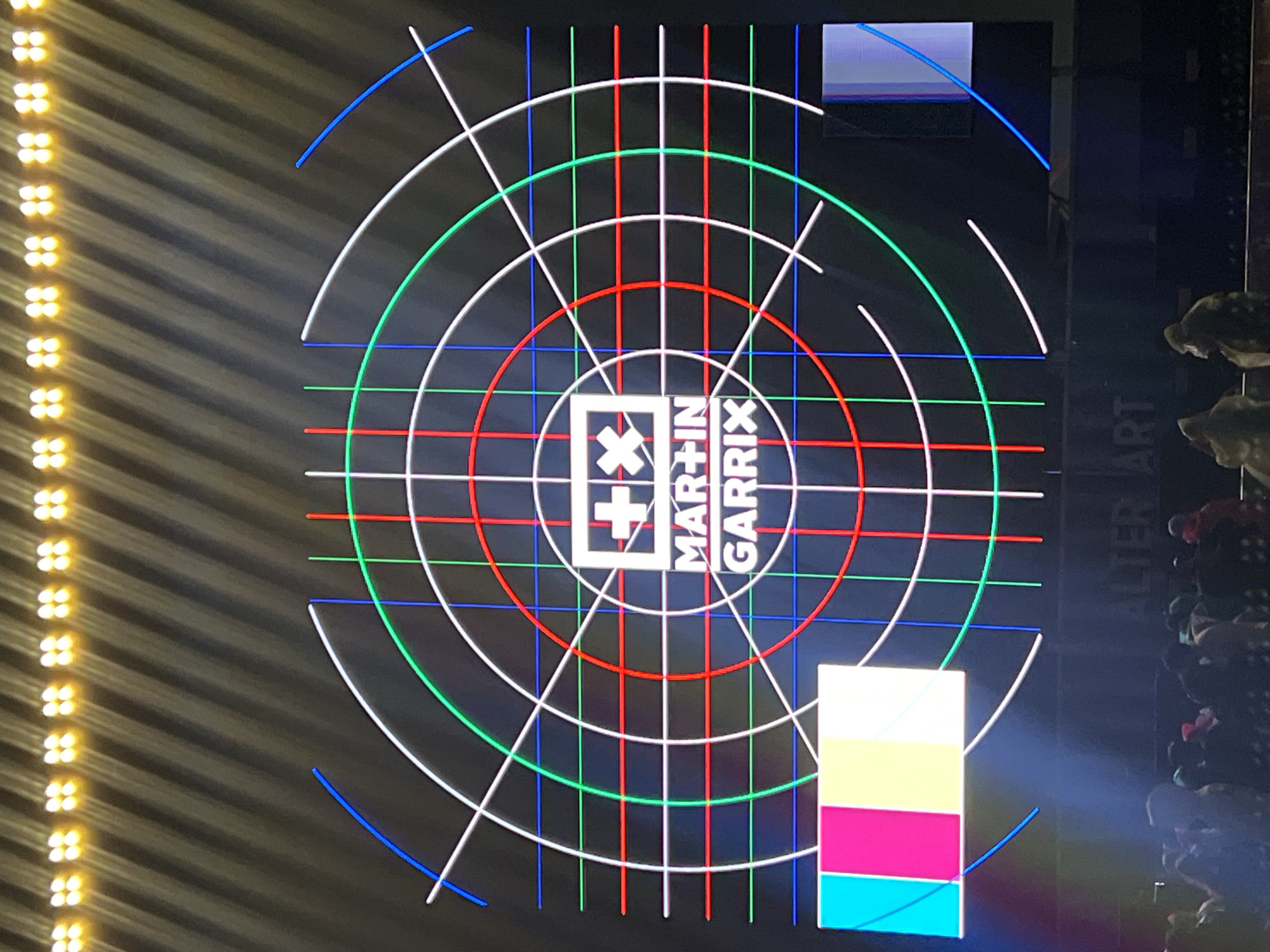 The image shows a large screen displaying a graphic with concentric circles and intersecting lines in blue, green, and red, reminiscent of a test pattern. In the center of the screen is a white logo consisting of a plus sign and an "X" within a square, above the text "MAR+IN GARRIX". Below the screen, a crowd of people is visible, silhouetted against the bright display. The text "ALTER ART" is visible at the bottom of the screen. The scene appears to be an outdoor concert or event.
Based on the available information, Martin Garrix performed at Alter Art Festival in Gdynia, Poland. Therefore, the location is likely the Alter Art Festival grounds in Gdynia, Poland.