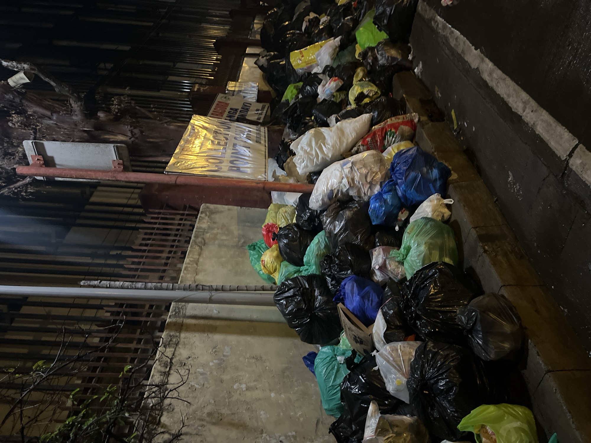 The image depicts a large accumulation of garbage bags overflowing onto the sidewalk in Davao City, Philippines. The bags are various colors, including black, green, blue, white, and yellow. A sign is partially visible behind some of the bags, with the words "COLLECTION POINT BASURA" and "MGA LUNES HANGGANG LINGGO" written on it. The area appears to be next to a building with a metal fence. The lighting suggests it is night. The sidewalk is made of concrete.