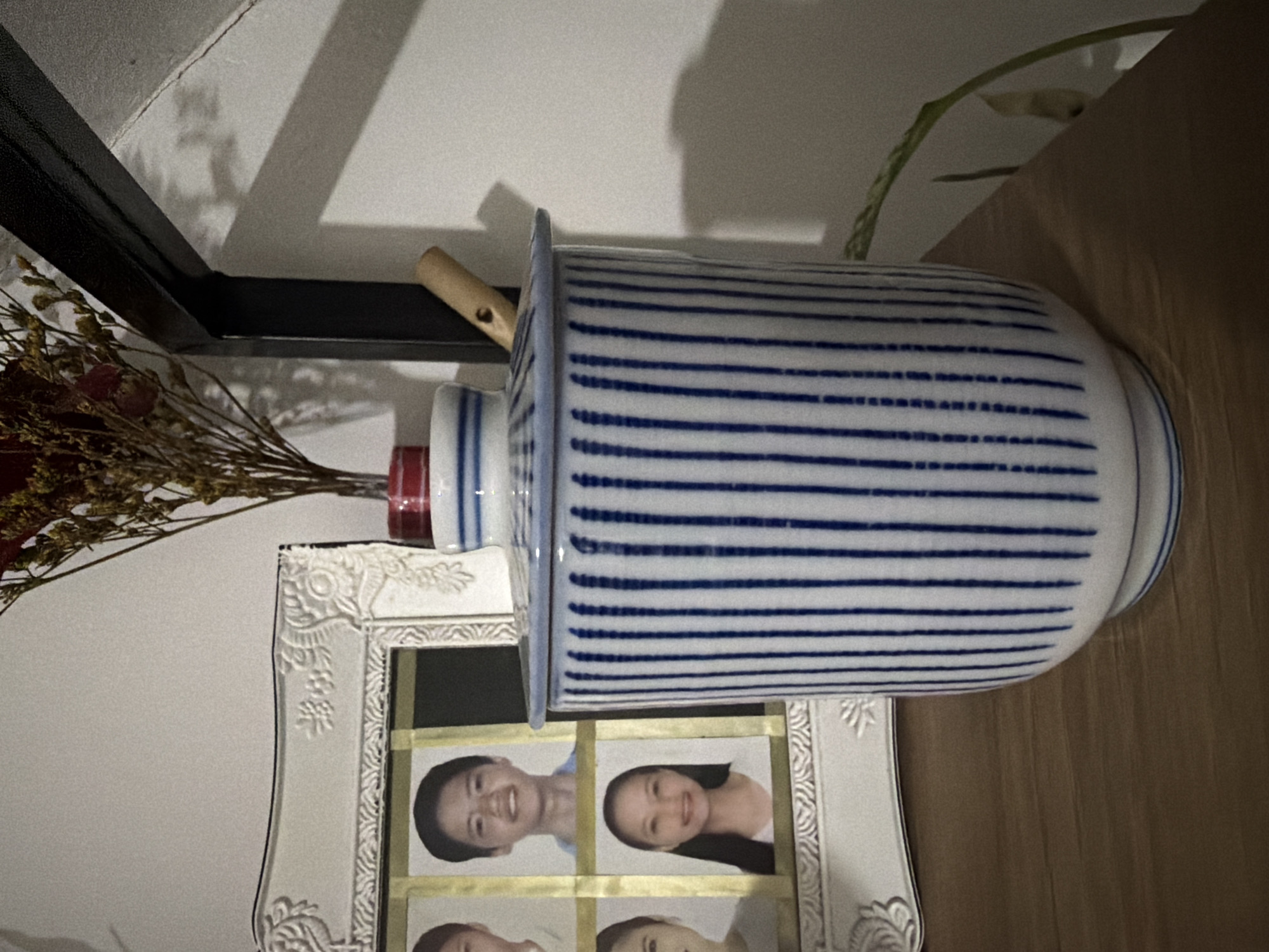 A blue and white striped ceramic jar with a wooden spoon rests on a wooden surface. Behind the jar, a framed photo displays four passport-sized portraits of individuals. To the right of the jar, a dried floral arrangement in a red and white vase is visible. The background appears to be a wall with shadows cast from objects. There is no indication of the specific location within Davao City, Philippines.