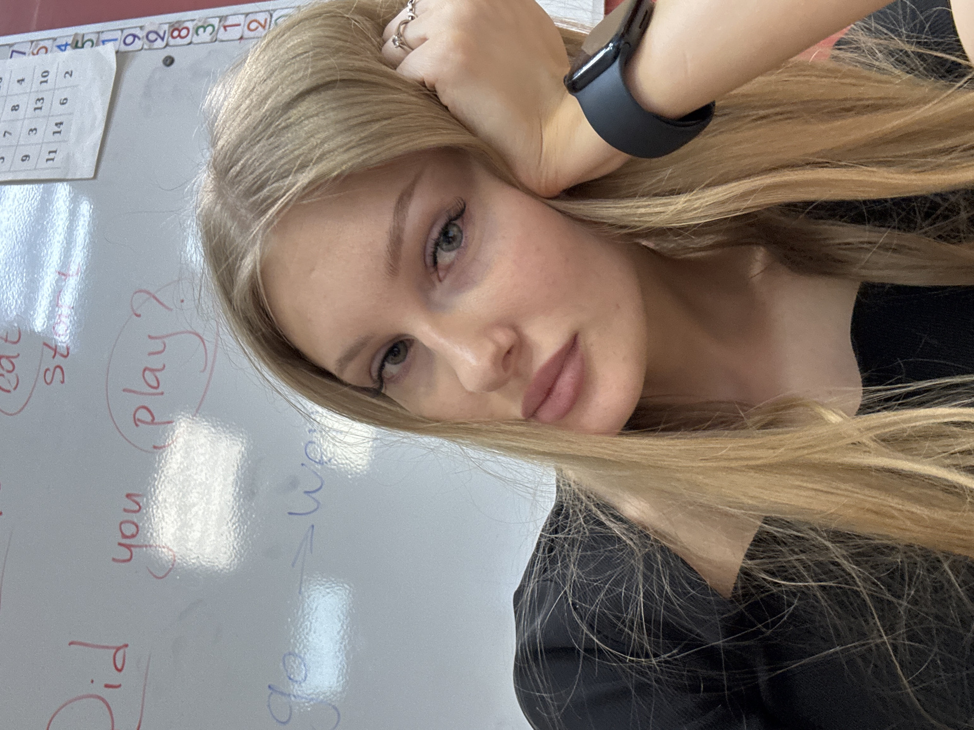 A young woman with blonde hair is posing for a selfie. Her left arm is bent, with her hand resting on her head, wearing a black smartwatch on her wrist. She has light skin, green eyes, and pink lips. She is wearing a black top. In the background is a white board with the words "Did you play?" and "go -> We" written in red and blue marker. A calendar is visible in the upper left corner of the whiteboard with the numbers 1-14.