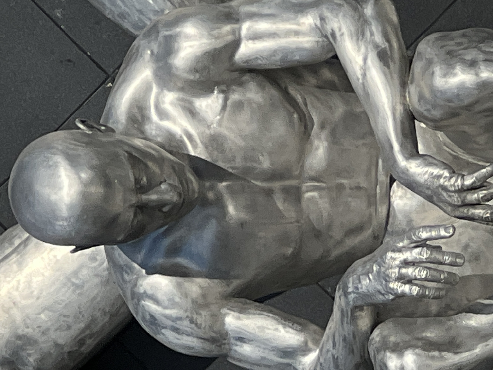 This is a close-up, eye-level, medium shot of a metallic sculpture of a seated, bald man. The sculpture is a realistic depiction of a muscular male physique, with visible abdominal muscles and defined arms. The man is looking downwards, with his head bowed and his hands resting on his knees. The sculpture has a highly polished, reflective surface, giving it a silver appearance. It is situated outdoors, with a dark, tiled wall visible in the background. The lighting suggests it is daytime, with strong highlights on the sculpture. This sculpture is located at the entrance to the "Park of the Year 2000" in Krasnodar, Russia.
