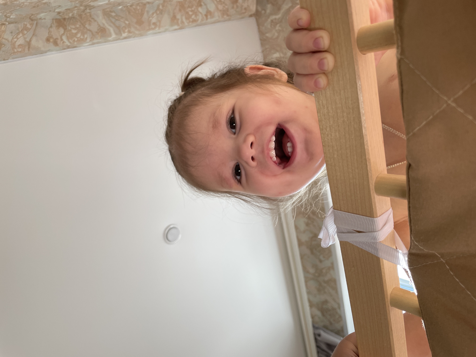 A young child with brown hair pulled back in a small ponytail is looking down and smiling with her mouth open, revealing her teeth. The child is holding onto a light-colored wooden structure with her right hand. A white ribbon is tied around the wooden structure. In the background, there is a white wall with a light fixture and wallpaper with a light brown and white pattern. The image was likely taken indoors. There is no information available from the image to confirm that it was taken in Izhevsk, Russia.