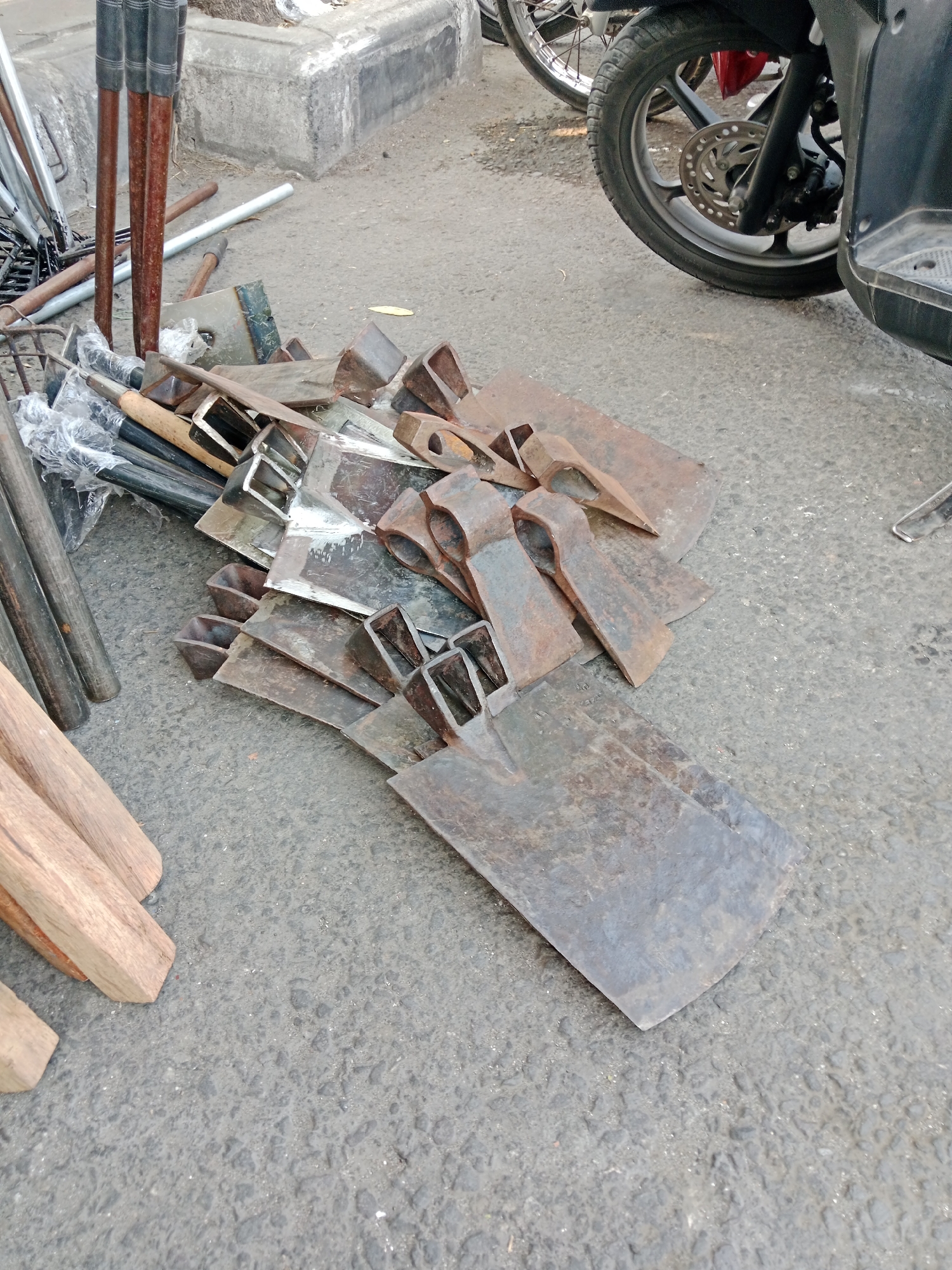 A pile of farm tools, including hoes and axes, rests on an asphalt street in Kediri City, Indonesia. The tools are made of metal and wood and show signs of rust and wear. In the background, a motorcycle is parked, and a concrete curb is visible. The scene appears to be an outdoor market or a roadside stall.