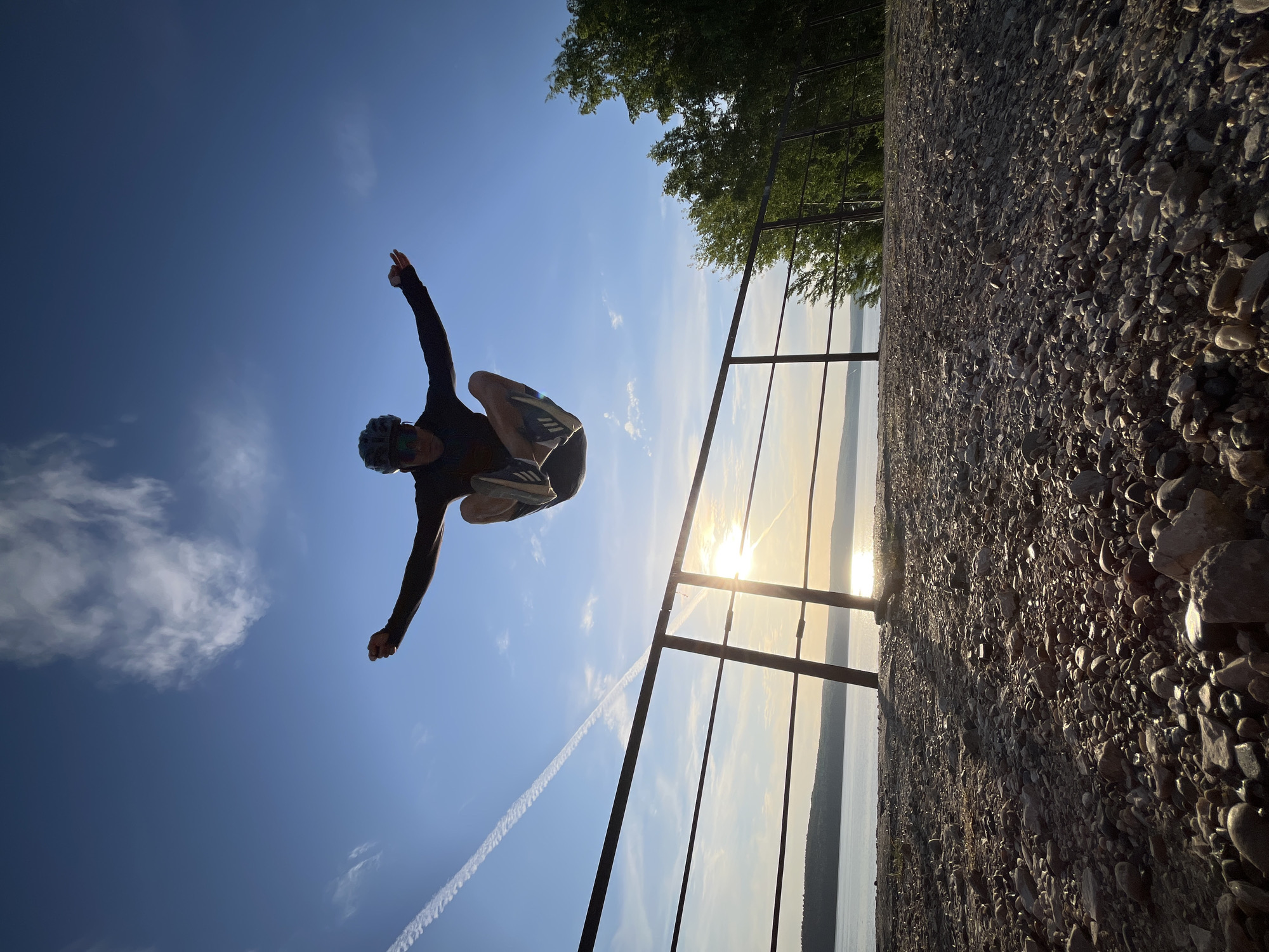 A low-angle shot depicts a man in mid-air against a blue sky. He is wearing a helmet and dark long-sleeved shirt with light shorts and athletic shoes. His arms are outstretched. In the background is a view of water and trees obscured by a metal railing and a rocky shoreline. The sun is reflecting off the water, positioned directly behind the railing. A vapor trail is visible in the sky. Based on the visual evidence, this location is likely in or near Chaikovsky, Russia, considering the prompt specifies this city. The man appears to be performing a jump or gymnastic maneuver.