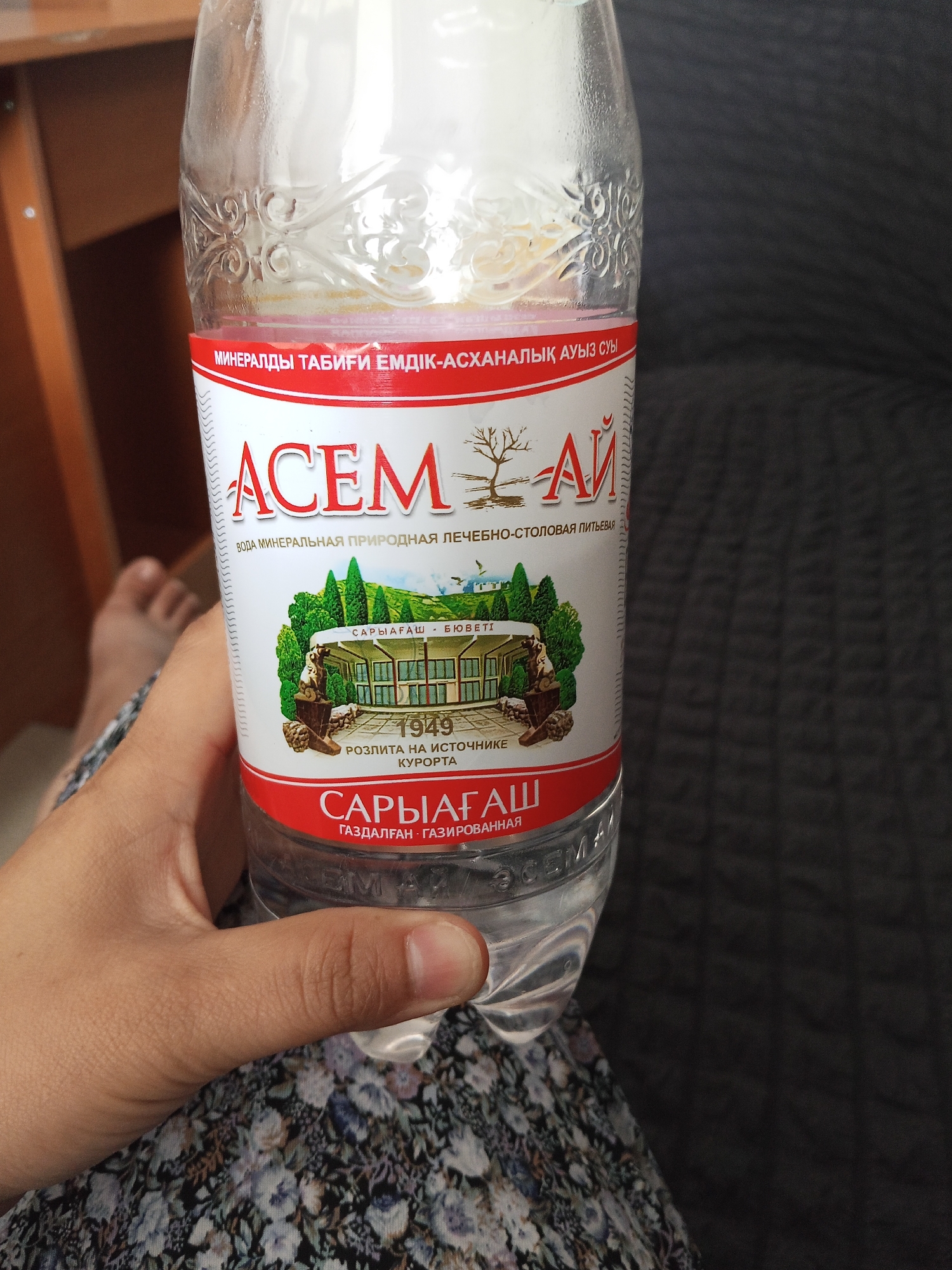 A person's hand holds a clear plastic bottle of mineral water. The bottle has a prominent red and white label with text in both Cyrillic and Latin scripts, indicating it is "ACEM" brand mineral water from "Saryagash." The label also features an illustration of a building surrounded by trees, along with the year "1949" and text in Russian stating it is "Bottled at the source of the resort." The background is blurred, showing a wooden surface and a dark gray fabric, suggesting an indoor setting. The person's bare foot is partially visible in the lower left corner, resting on a patterned fabric. The image appears to be taken during daylight hours.