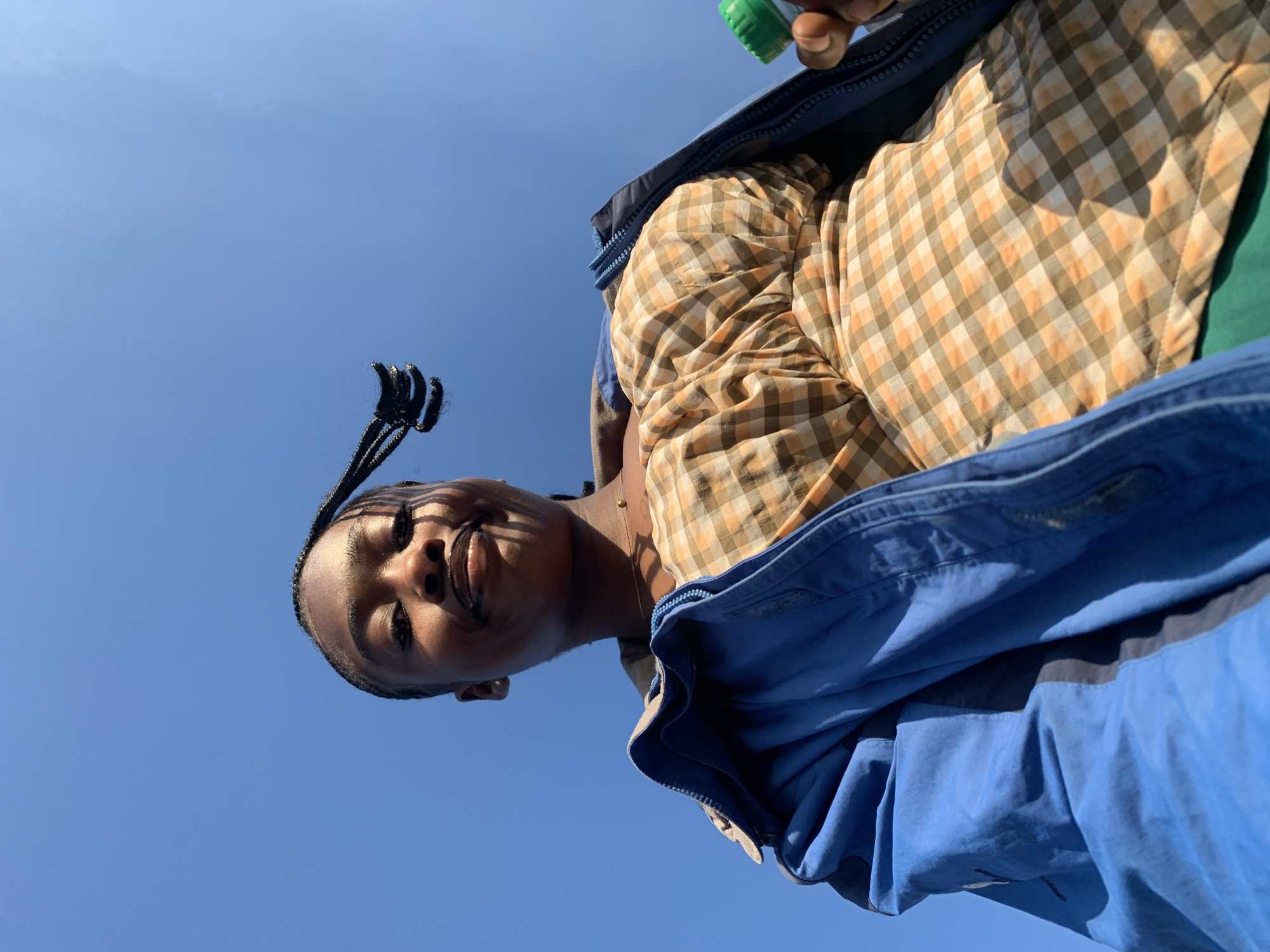 A low-angle, eye-level, medium shot captures a young Black woman from the shoulders up. She is looking up at the camera with a slight smile and her eyes are closed. Her dark hair is styled in braids, with one braid extending up and away from her head. She is wearing a light brown and white checkered shirt underneath a blue jacket. The blue jacket is partially unzipped, revealing the checkered shirt. Her right hand is visible on the right side of the frame, holding a small green cap. The background is a clear, bright blue sky, with no visible clouds or distinguishing features of Rafin Par, Nigeria. The lighting suggests it is daytime, with sunlight casting shadows on the woman's face.