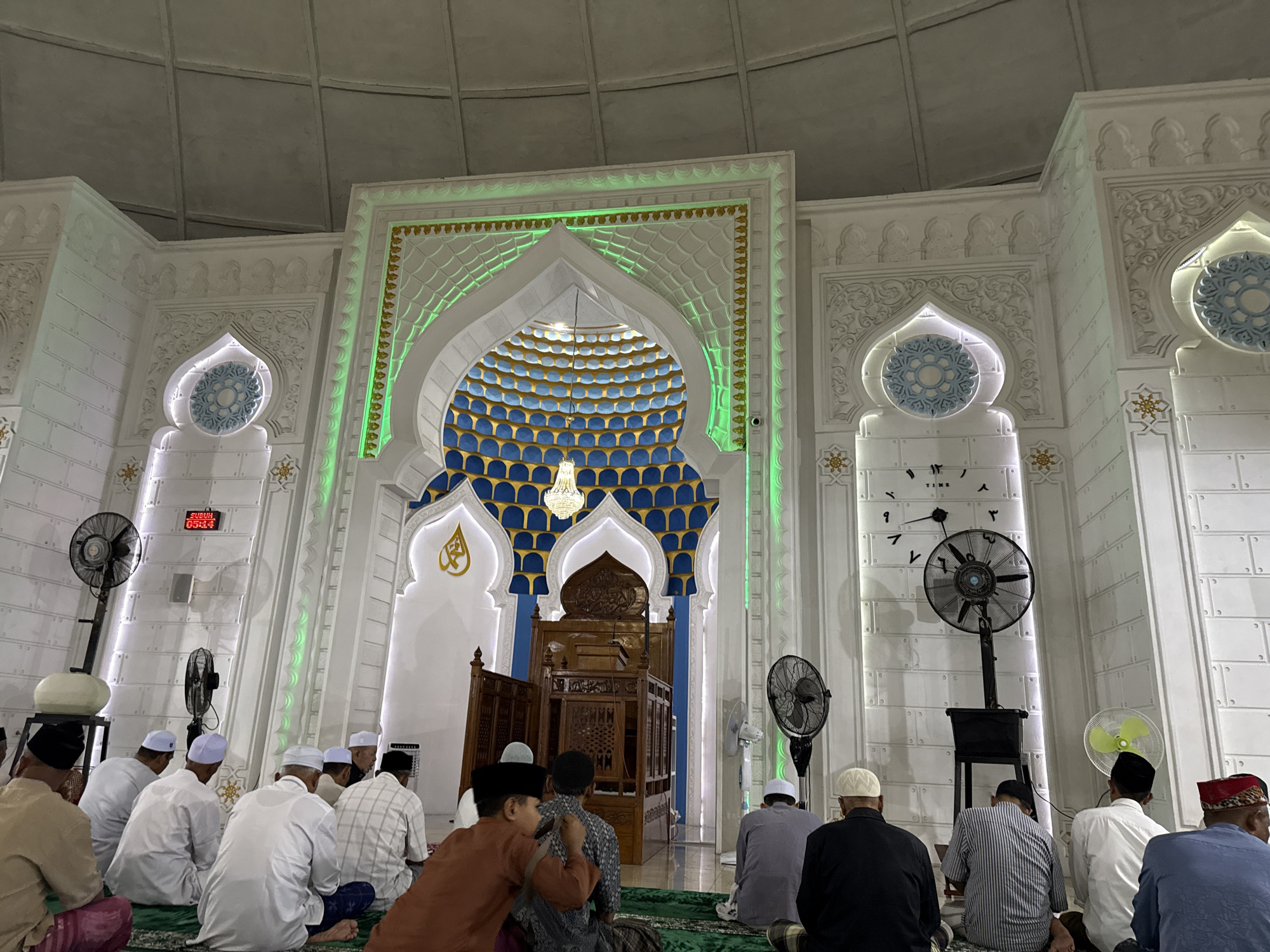 The image shows the interior of a mosque in Pasar Paloh, Indonesia, during prayers. A group of men, mostly wearing white skullcaps, are seated on a green carpet. Some are looking forward, while others are interacting with each other. The background features an elaborately decorated Mihrab with blue and gold patterns and green lighting. A wooden Minbar sits in front of the Mihrab. On the side walls, there are clock-like decorations and electric fans. A digital clock displays the time above the doorway on the left wall.