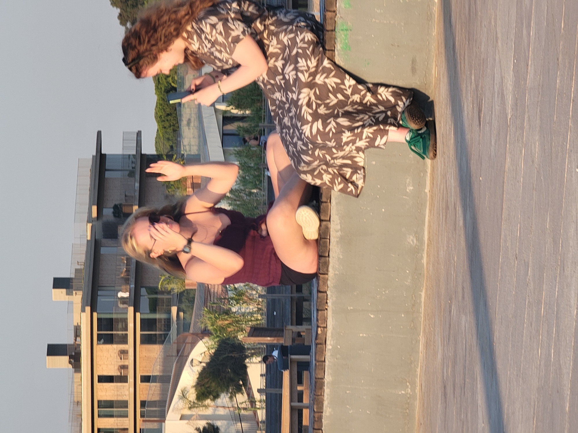 Two young women are seated on a low, weathered concrete wall in Quarteira, Portugal, with a building and trees in the background. The woman on the left, with blonde hair, wears a dark red tank top and black shorts. She is holding her hand up to her face, as if shielding her eyes or posing for a photo, and her left leg is bent with her foot resting on the wall. The woman on the right has long, curly brown hair and is wearing a patterned dress with a floral design. She is holding a smartphone in her hands, appearing to take a picture or video. She is wearing green sneakers. The wall they are sitting on is aged and has a textured surface. Behind them, a modern building with multiple balconies and large windows is visible, surrounded by lush green trees. The scene appears to be outdoors, possibly in a park or public space.