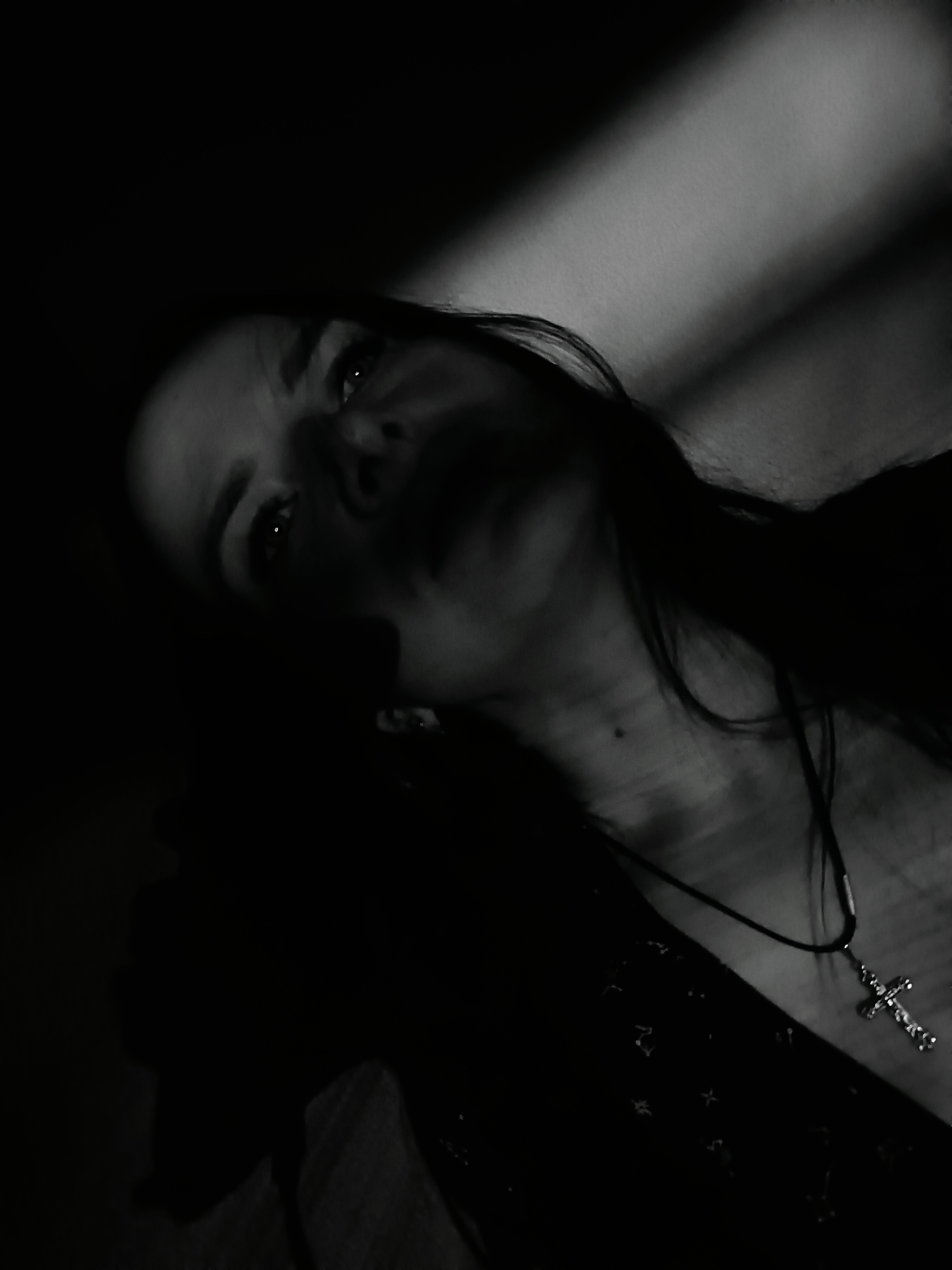 A low-angle, black-and-white photograph depicts a person with long hair, wearing a pendant with a cross, and gazing upwards. The lower portion of the face is obscured by shadow. The background features a lighter, smooth surface with an elongated streak of light. The location in the city of Rusalevka, Russia, cannot be confirmed based on the image alone.