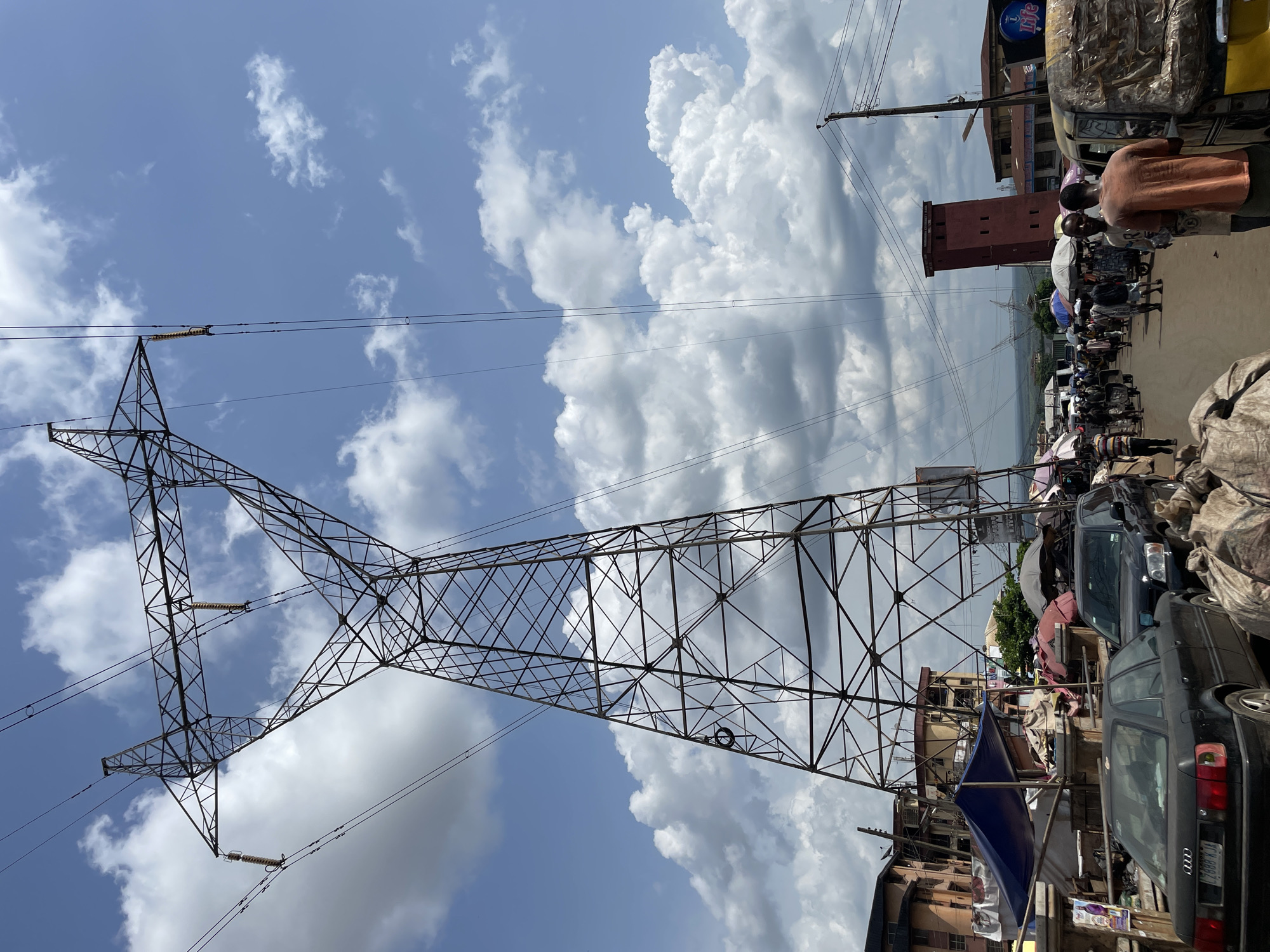 The image captures a vibrant street scene in Obosi, Nigeria, under a bright, cloudy sky. A massive metal lattice electricity pylon, extending from bottom to top, dominates the left side of the frame, showcasing its intricate structure and attached power lines.
On the right, a bustling commercial or residential street unfolds, populated by numerous individuals, vehicles, and buildings. Several people are visible, some standing, possibly engaged in market activities, with two individuals prominently in the mid-ground. Parked cars, including a dark Audi sedan with visible branding and a partial license plate, line the street, alongside part of what appears to be a tricycle taxi.
The street is characterized by temporary market stalls, goods, and various covers, including a prominent blue tarpaulin. In the background, a mix of buildings can be seen, notably a distinctive red-brown, tower-like structure with small rectangular windows. Overhead, the sky is a vivid blue, filled with large, white, fluffy cumulus clouds, indicating fair daytime weather. A blue sign with the word "Life" is partially visible in the background. The overall scene depicts a lively urban environment with daily activities taking place.