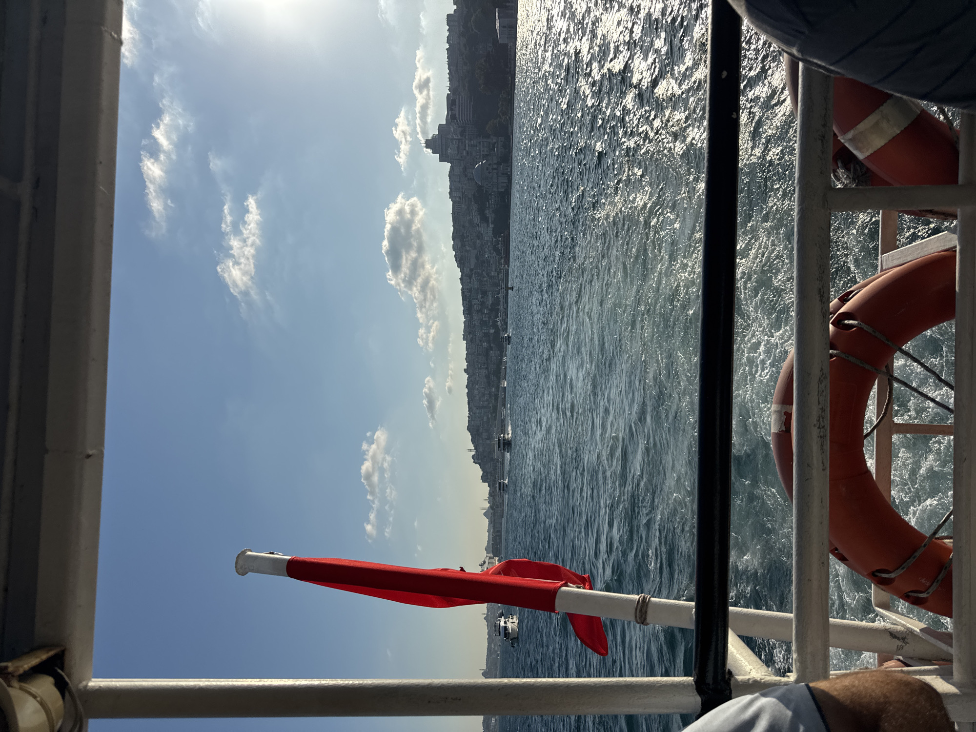 This is a medium shot from a boat traveling on the Bosphorus Strait, with the city of Istanbul, specifically the Beşiktaş district, visible in the distance on the European side. The foreground features the stern of the boat, with a red flag attached to a pole on the left side and orange life preservers mounted on the railing. Two people are partially visible, one on the far left and another on the far right, seated on the boat. The water is choppy, creating white wakes behind the boat. The sky is blue with scattered white clouds and the sun is shining brightly, reflecting off the water. The cityscape of Istanbul features buildings of various sizes and architectural styles, including domes and minarets, indicating a significant urban area. Several other boats are seen on the water, some in the middle distance and some further away. The overall scene depicts a sunny day trip on the Bosphorus.