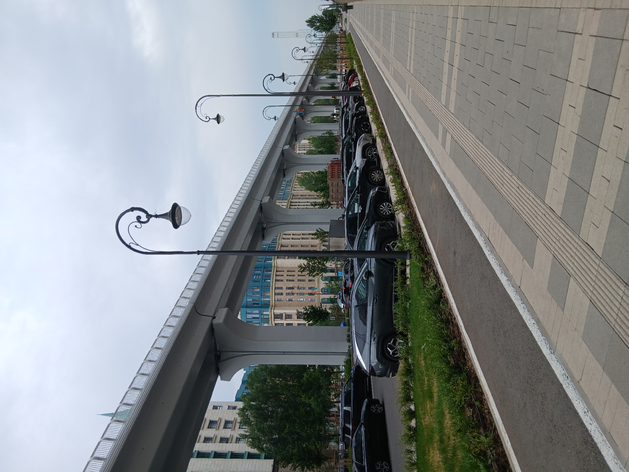The image captures a street scene in Astana, Kazakhstan, featuring a modern elevated roadway or bridge structure that spans across the frame from left to right. Beneath the elevated structure, a row of parked cars lines a street, with a pedestrian walkway adjacent to the parking area. The walkway is paved with light-colored rectangular tiles and features a tactile paving strip for visually impaired individuals. Several ornate street lamps with curved arms and glass fixtures are visible along the pedestrian path, punctuating the urban landscape. In the background, contemporary buildings with light-colored facades and some with blue accents rise, indicating a developed urban environment. The sky is overcast with clouds, suggesting a diffused light condition. The scene is devoid of people, and no specific actions or interactions are depicted. The overall impression is one of urban infrastructure and public space under a cloudy sky.