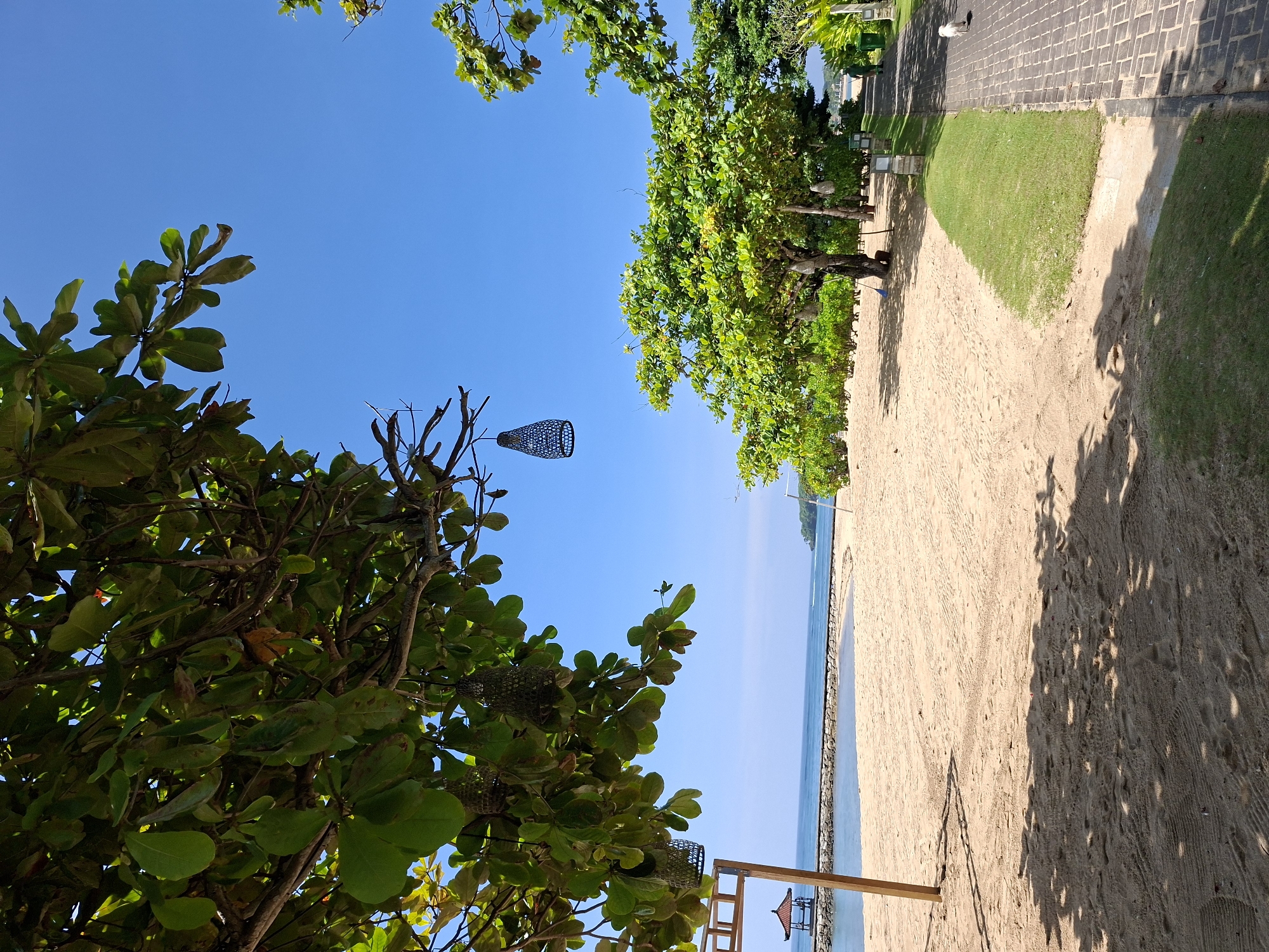 The image displays a bright, sunny beach scene, likely situated in Kuta Selatan, Indonesia. The perspective is from under a canopy of lush green tropical trees, looking towards the ocean.
In the foreground and along the left side, vibrant green leaves and branches of several trees frame the view. Among the branches, multiple decorative, dark-colored mesh lanterns are suspended. The sky above is a clear, brilliant blue, indicating fair weather.
A wide stretch of light brown sand dominates the center and lower right of the image, showing subtle tracks and footprints. Shadows from the trees fall distinctly across the sand, suggesting a strong midday or early afternoon sun. The sandy beach transitions into the calm, blue ocean on the far right. A low stone wall or breakwater marks the edge between the sand and the water. Further out in the water, a small wooden structure, possibly a gazebo or part of a pier, is visible.
To the upper right, beyond the sandy area, a well-kept green lawn is visible, bordered by a paved walkway constructed from dark, rectangular tiles. Additional trees line this path, contributing to the lush environment.
The scene is devoid of people, conveying a tranquil and serene atmosphere. The bright sunshine, deep blue sky, and strong shadows all point to a pleasant day. No text is visible within the image.