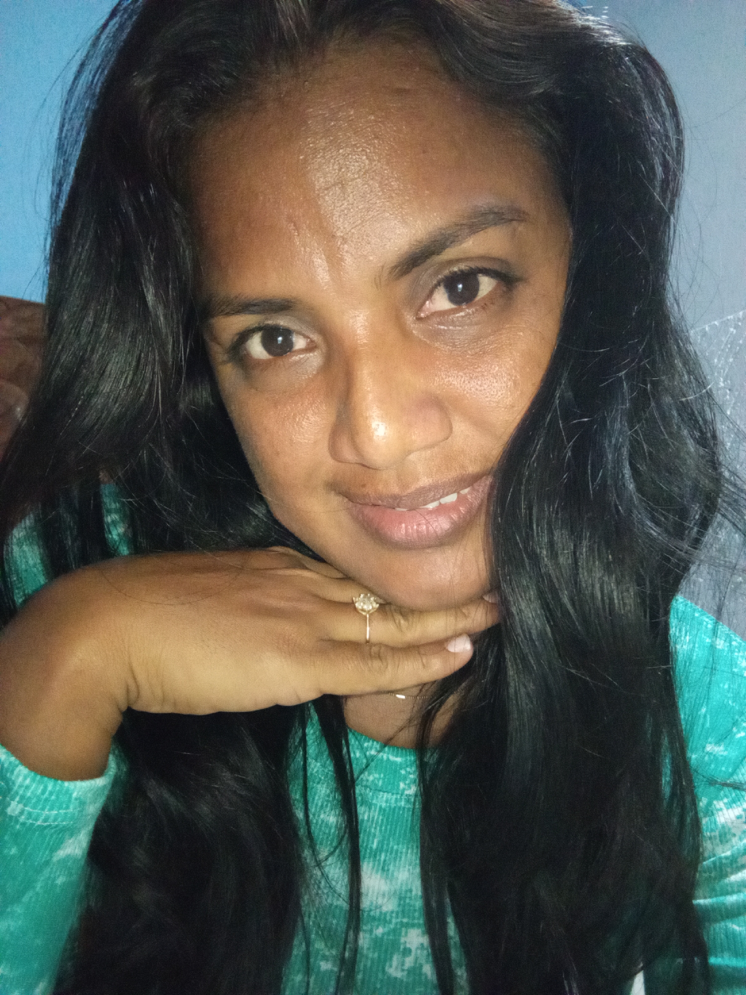 The image is a close-up, waist-up portrait of a woman. She has dark, long hair that frames her face and falls over her shoulders. Her skin is a medium brown tone, and she has dark eyes. She is wearing a turquoise shirt with a textured pattern. Her right hand is resting on her chin, and she is wearing a ring with a diamond-like stone on her ring finger. The background is out of focus, with a hint of a blue wall on the left and a gray surface on the right. The lighting is somewhat dim, with a glow highlighting her forehead and cheeks. The woman is looking directly at the camera with a slight smile.