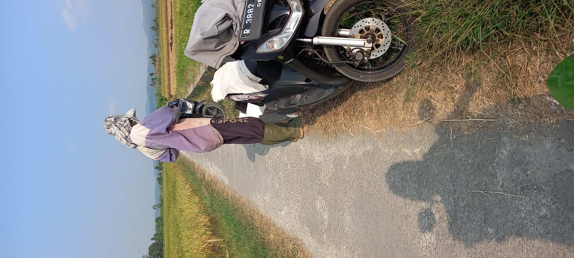 In Sidamulya, Indonesia, on a paved road bordered by a field of golden rice and dry grass, two figures are visible near parked motorcycles under a clear blue sky. The figure in the foreground, facing away from the camera, is wearing dark pants with white lettering, green rubber boots, and a two-toned jacket with purple and cream panels, topped with a camouflage patterned hat. This individual stands beside a black motorcycle covered with a grey cloth, and a white cloth is draped over the handlebars. Another motorcycle is parked behind it. A shadow of a person, presumably the photographer, is cast on the road to the left. The background shows more rice fields and distant trees and hills.