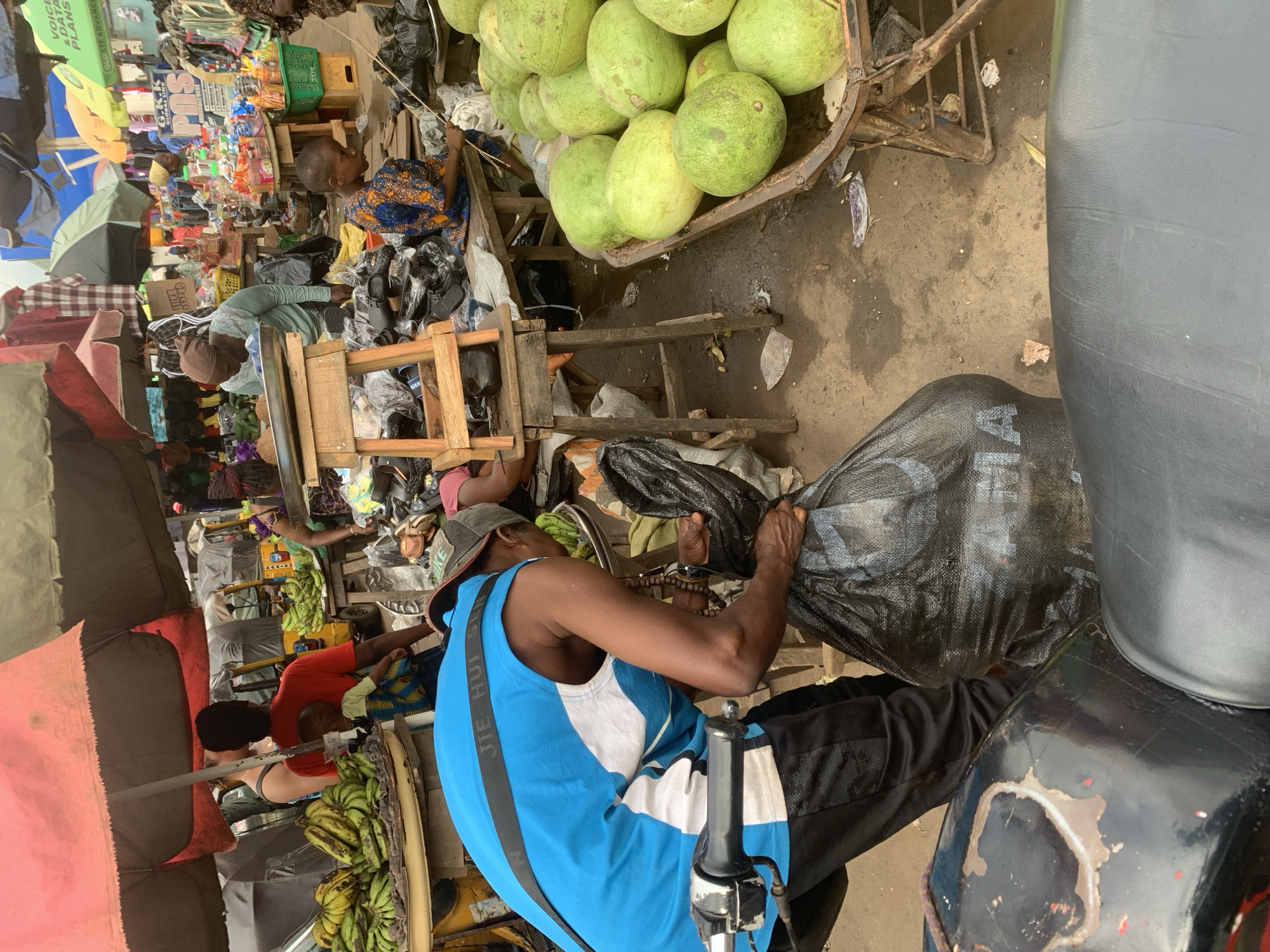 The image captures a bustling outdoor market scene, likely in Shagamu, Nigeria. The foreground is dominated by a person wearing a blue and white sleeveless shirt and black pants, stooping to lift a large black plastic bag. A motorcycle, partially visible in the lower portion of the frame, suggests this person may be involved in transport.
Behind the person, the market unfolds with various stalls and activities. Bunches of green bananas are piled high on a tray. Various goods are displayed on wooden tables and stands, including what appear to be watermelons in a metal cart on the right side of the image. Stalls are covered by colorful umbrellas and awnings in shades of red and green, providing shade from the sun.
Several other individuals are present in the market. A woman wearing a brown headscarf and a light blue top is seen interacting with the goods on a table. A child in a blue and orange patterned outfit is seated on a wooden bench. Other figures are visible in the background, engaged in market activities. The ground is dirt and appears damp in places. The overall impression is one of a vibrant and active marketplace.