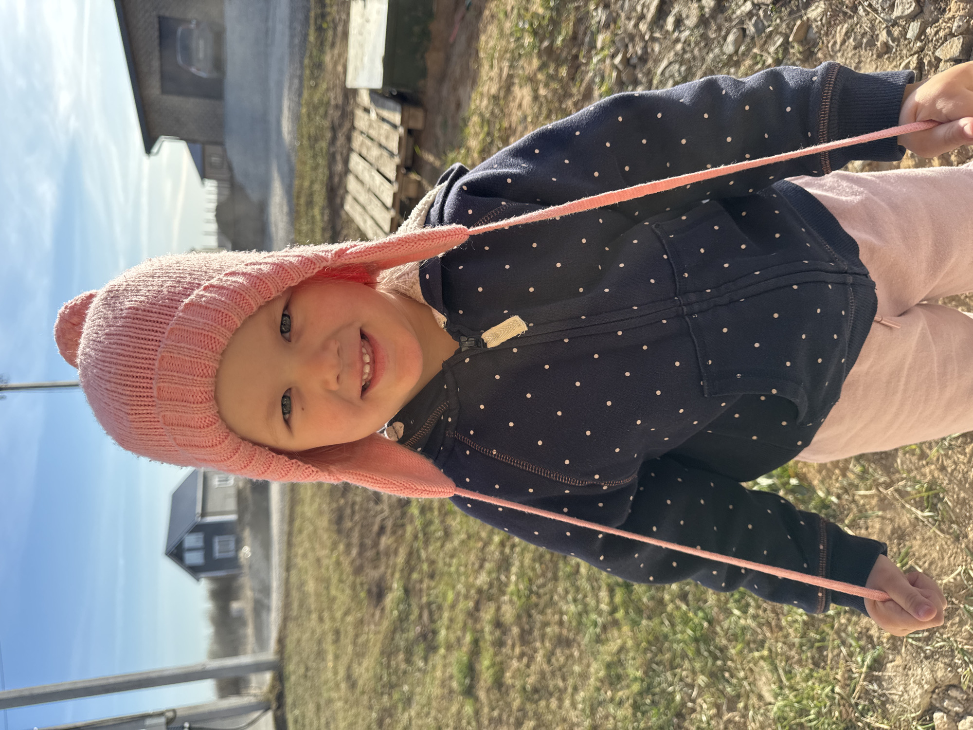 A cheerful young child, likely a toddler, is depicted outdoors in Gornyi (Dvurechenskoe S/P), Russia, on a bright, sunny day. The child, with light skin and blue eyes, is looking directly at the camera with a wide, open-mouthed smile, revealing some teeth, indicating happiness and playfulness. They are wearing a pink knitted hat with ear flaps and small pom-poms on top, a dark blue hooded zip-up jacket adorned with white polka dots, and light pink pants.
The child is actively engaging with their hat, holding one of the pink strings attached to the ear flaps in each hand and pulling them outwards. The background suggests a rural or semi-rural setting with patches of dry grass and dirt. Distant buildings are visible under a clear, bright blue sky with some faint clouds, including a dark grey house with white window frames and another lighter structure. A wooden pallet or similar structure is seen on the right, and a metal railing is partially visible on the left side of the frame. The strong sunlight casts shadows, suggesting it is daytime, possibly late morning or afternoon. No legible text is visible on the child's clothing or in the background, though a small fabric tag is present on the jacket.