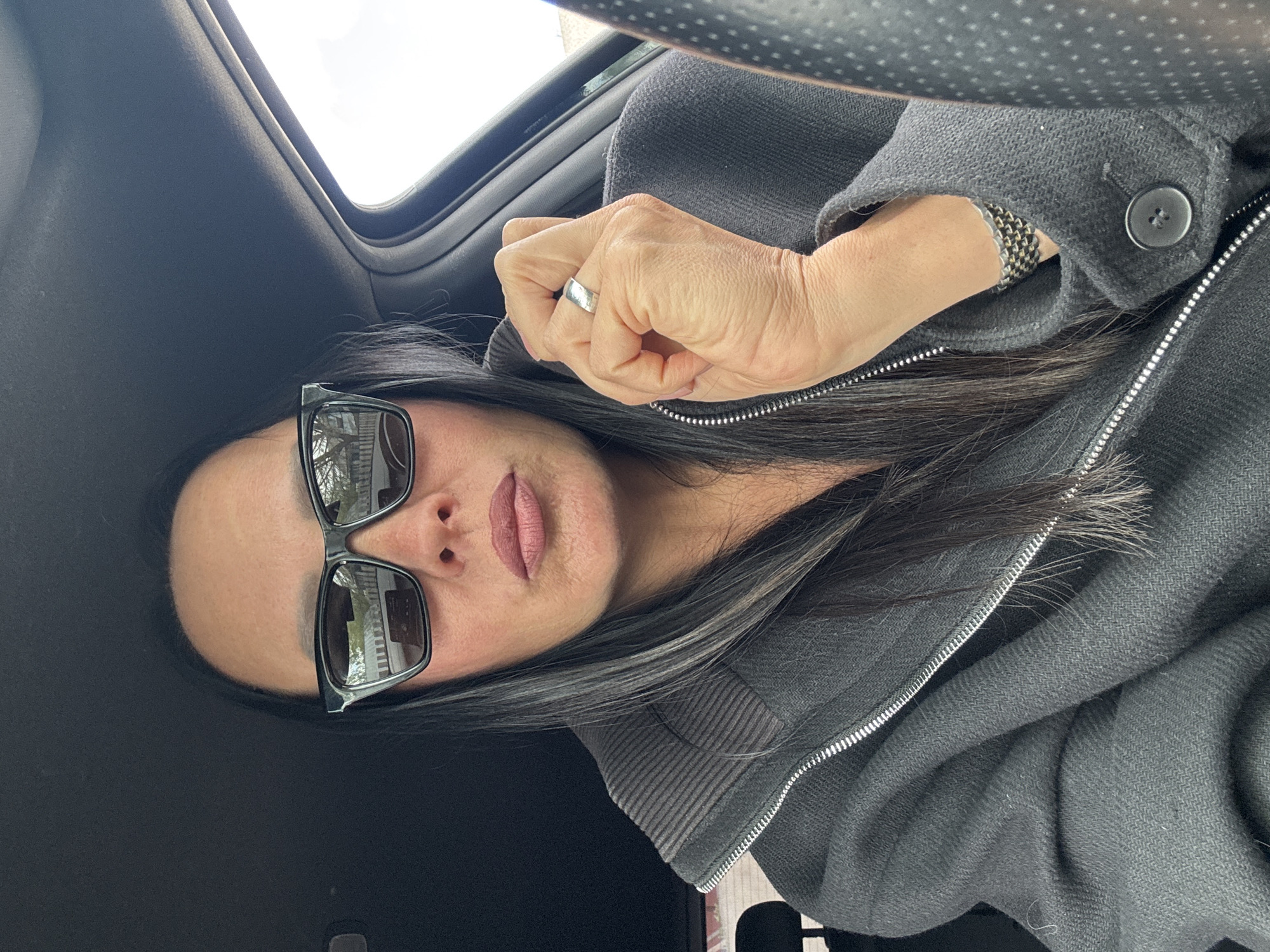 A woman wearing sunglasses is seated inside a vehicle. The vehicle's interior is dark. The woman is wearing a gray zippered jacket and has dark hair with gray streaks. She is wearing a ring on her left hand and a bracelet on her right. A sunroof is visible in the upper left. The location in the city of Chaikovsky, Russia, cannot be ascertained from the image.