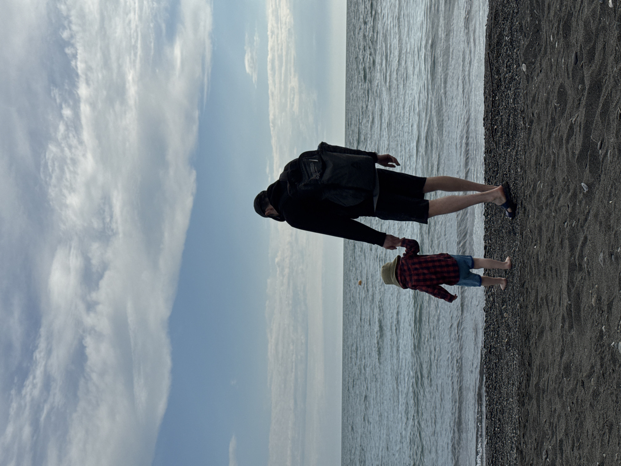 A father and a young child walk hand-in-hand on a beach in Sochi, Russia. The adult is wearing dark clothing, a backpack, and sandals, while the child wears a straw hat and a red plaid shirt. The beach has dark sand, and the sea meets the sky with a horizon line. White, wispy clouds fill the sky.