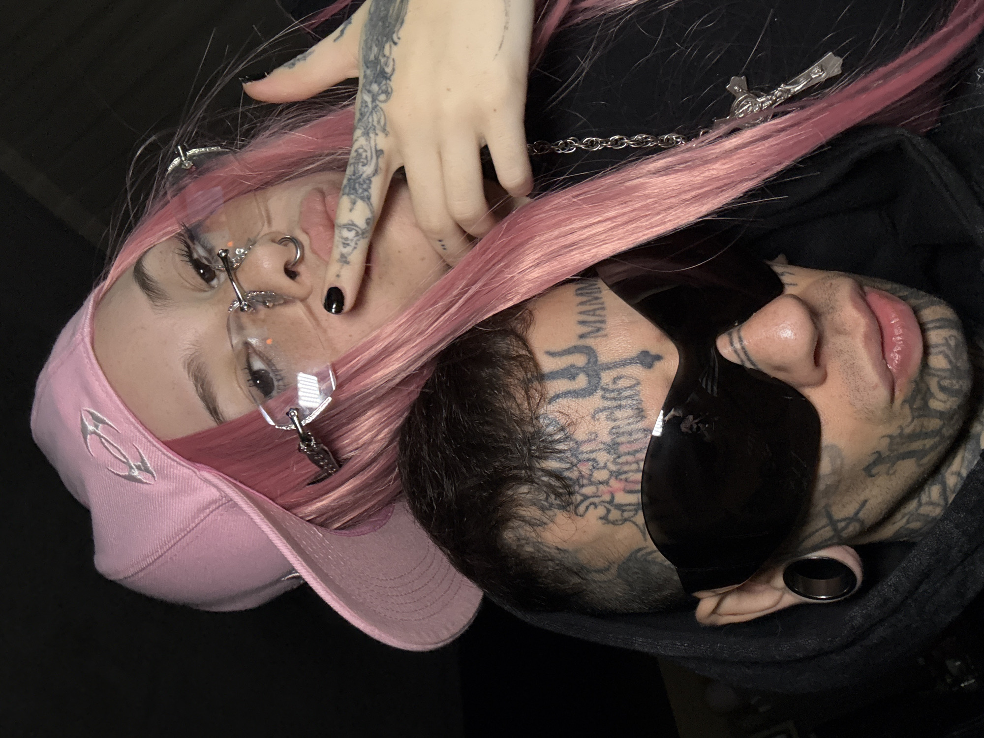 A close-up portrait features two individuals against a dark, indistinct background. A female-presenting individual, positioned top-right, has long, straight, vibrant pink hair and wears a light pink baseball cap with a silver-colored metallic emblem, worn backward. She wears rimless eyeglasses with complex silver metal frames, a small septum piercing, and black nail polish. Her left hand, adorned with intricate black tattoos, touches her pouting lips. A silver chain necklace with a crucifix pendant is visible on her right. A male-presenting individual, positioned bottom-left, displays extensive black tattoos covering his face, neck, and head, including script and symbols like a trident and the word "MAMMA." He wears large, dark, wrap-around sunglasses and a black ear gauge in his left ear. He is dressed in a dark hooded sweatshirt, with his lips slightly parted. The individuals are positioned intimately, suggesting a posed shot. The dark background offers no discernible features for location identification. The image provides no visual information to ascertain a specific location within Buenos Aires, Argentina.