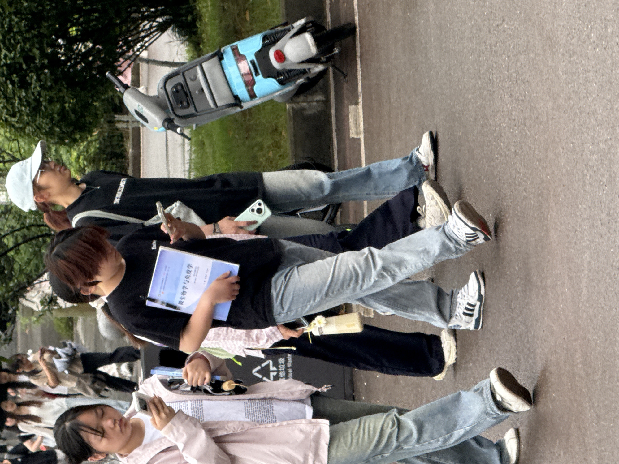 The image shows a group of at least 8 people standing on a paved area, likely a sidewalk, in the city of Jiangjin, China. A moped is parked on the sidewalk next to some grass and trees. The people are primarily young, and many are looking at or using mobile phones. One person is holding a book with text in Chinese, possibly related to microbiology and immunology. Some carry drinks. There is also a garbage bin present. The location appears to be a street or a public space.