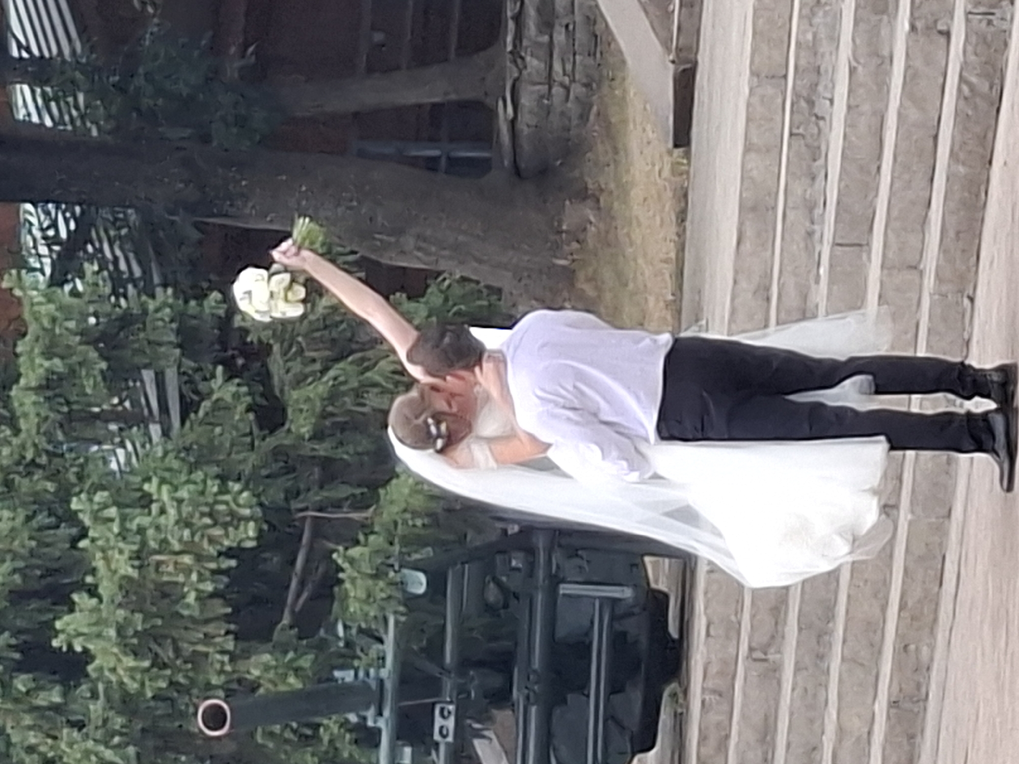A newly married couple, the bride in a white wedding dress and veil and the groom in a white dress shirt and black pants, are embracing on a stone staircase. The groom is lifting the bride, who is holding a bouquet of white flowers. In the background, a large green tree and what appears to be a military artifact, possibly an anti-aircraft gun, are visible. The scene is outdoors, with steps leading up to a building or structure behind the couple. This location is consistent with areas in Almaty, Kazakhstan, where such monuments or historical sites might be found.