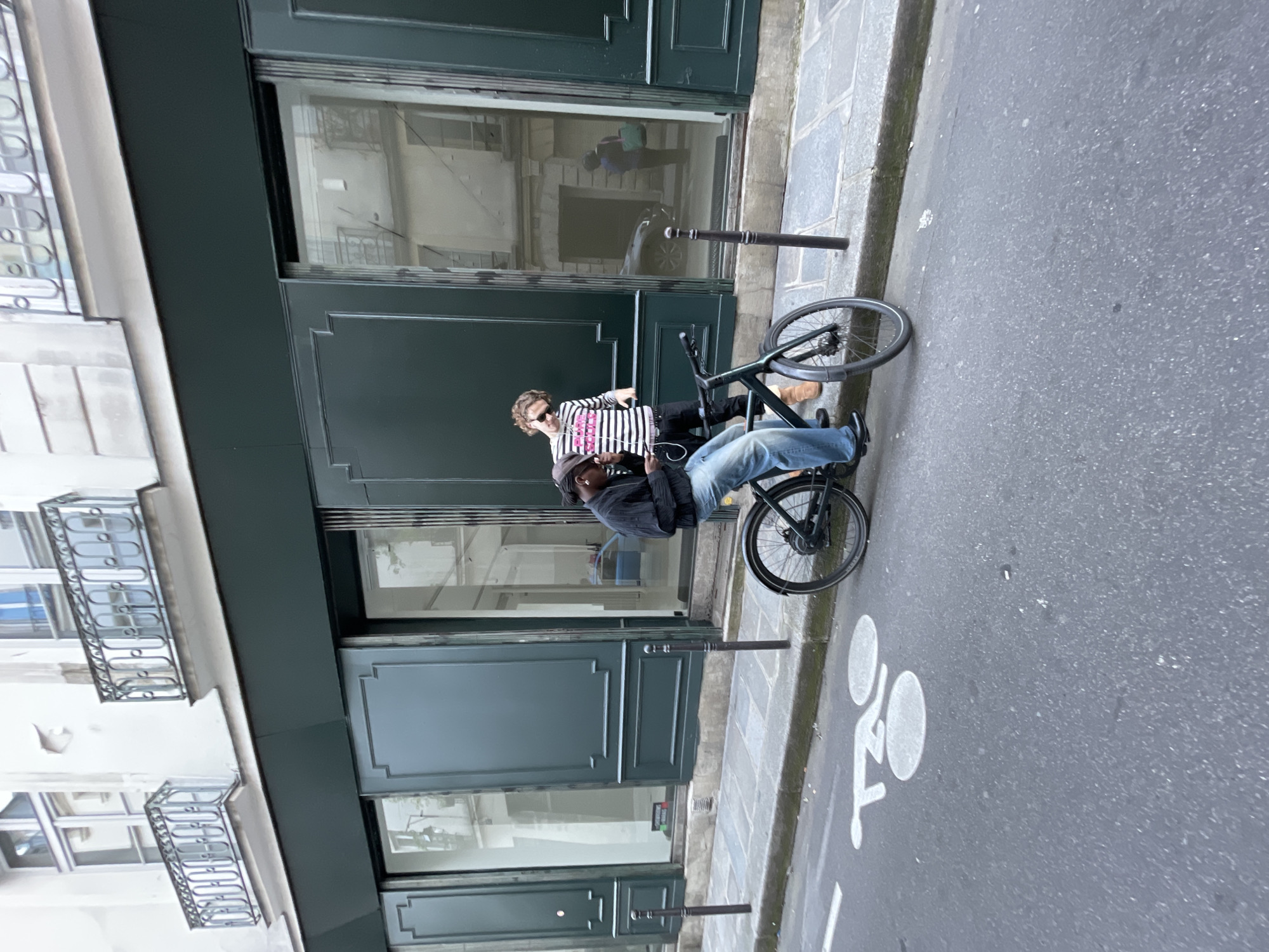 The image shows a street in Paris, France. The scene includes a building on the left with multiple floors and window balconies. The building has a storefront with large windows, and a dark green door. In front of the building, a man wearing a black jacket and jeans is leaning on a parked bicycle. A second individual, wearing a striped shirt and sunglasses, stands behind the bicycle. The road to the right features a white bicycle lane symbol painted on the surface. A metal pole, possibly a bike rack, is positioned between the building and the roadway. The overall scene appears to be a casual, everyday moment on a Parisian street.