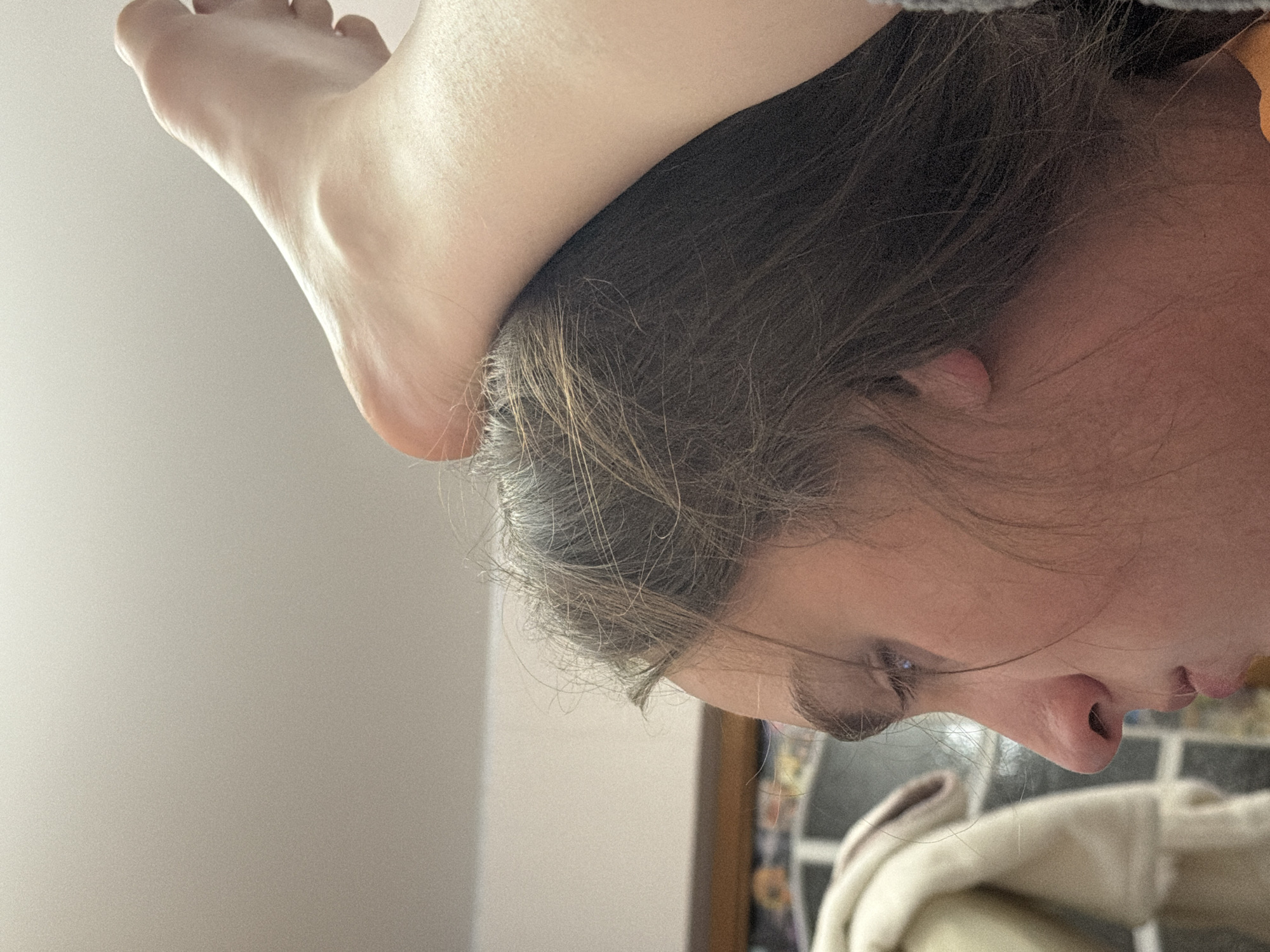 A person's foot rests on the head of a person with brown hair. The face of the person is visible with eyes, nose, and mouth. The background is blurry but appears to be an indoor setting, possibly a home. There is no indication of the city of Perm, Russia.
