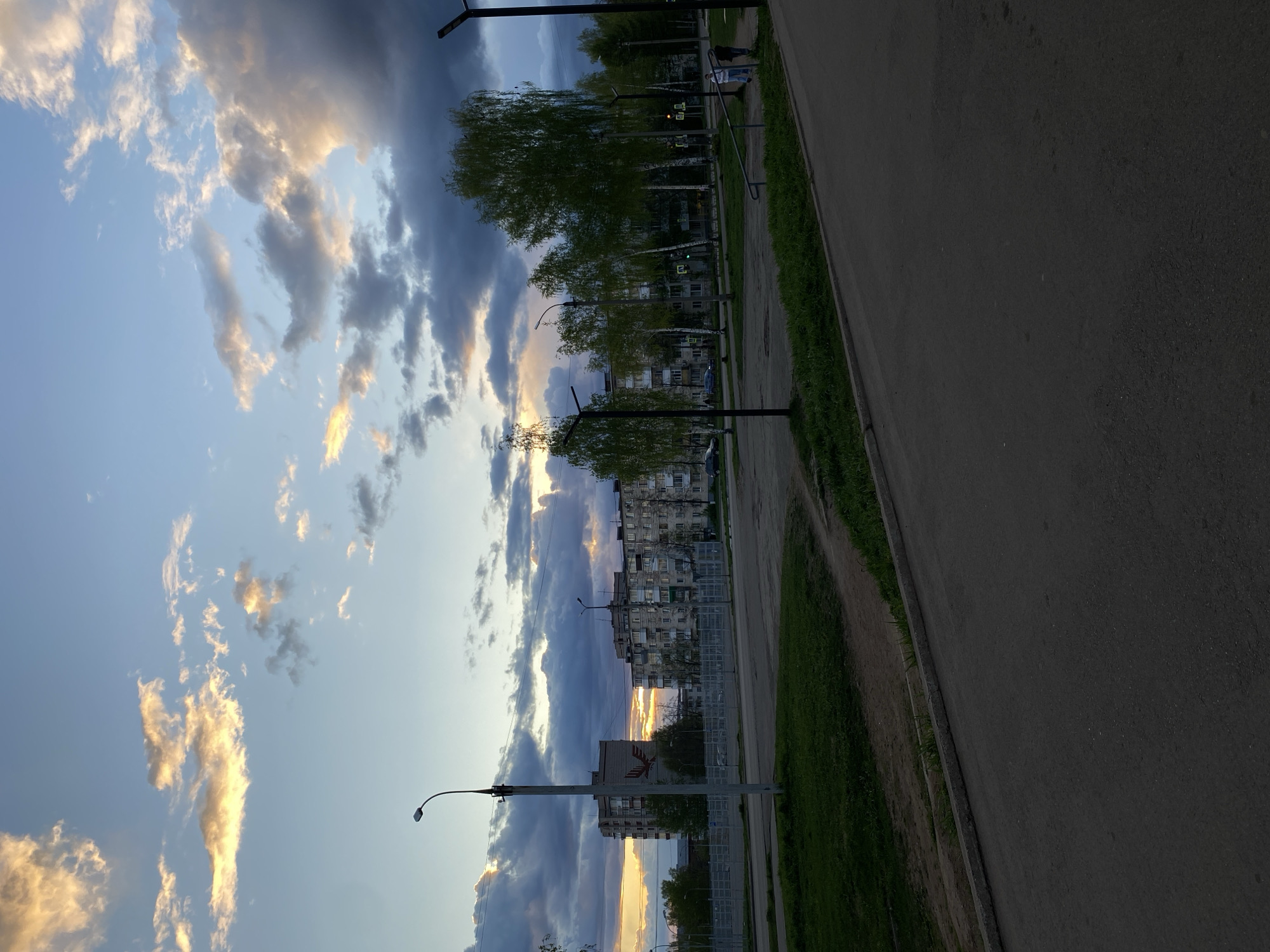 The image depicts an urban landscape at sunset in Votkinsk, Russia. The sky is filled with dynamic clouds, ranging from bright, sunlit cumulus to darker, ominous storm clouds. Sunlight filters through the clouds, creating a dramatic effect.
A row of trees lines the street, interspersed with streetlights. Buildings, likely residential apartments, are visible behind the trees. A paved road and a grassy area with a dirt path run along the right side of the image. A fence is also present in the image. There are a couple of people in the distance walking along the sidewalk.