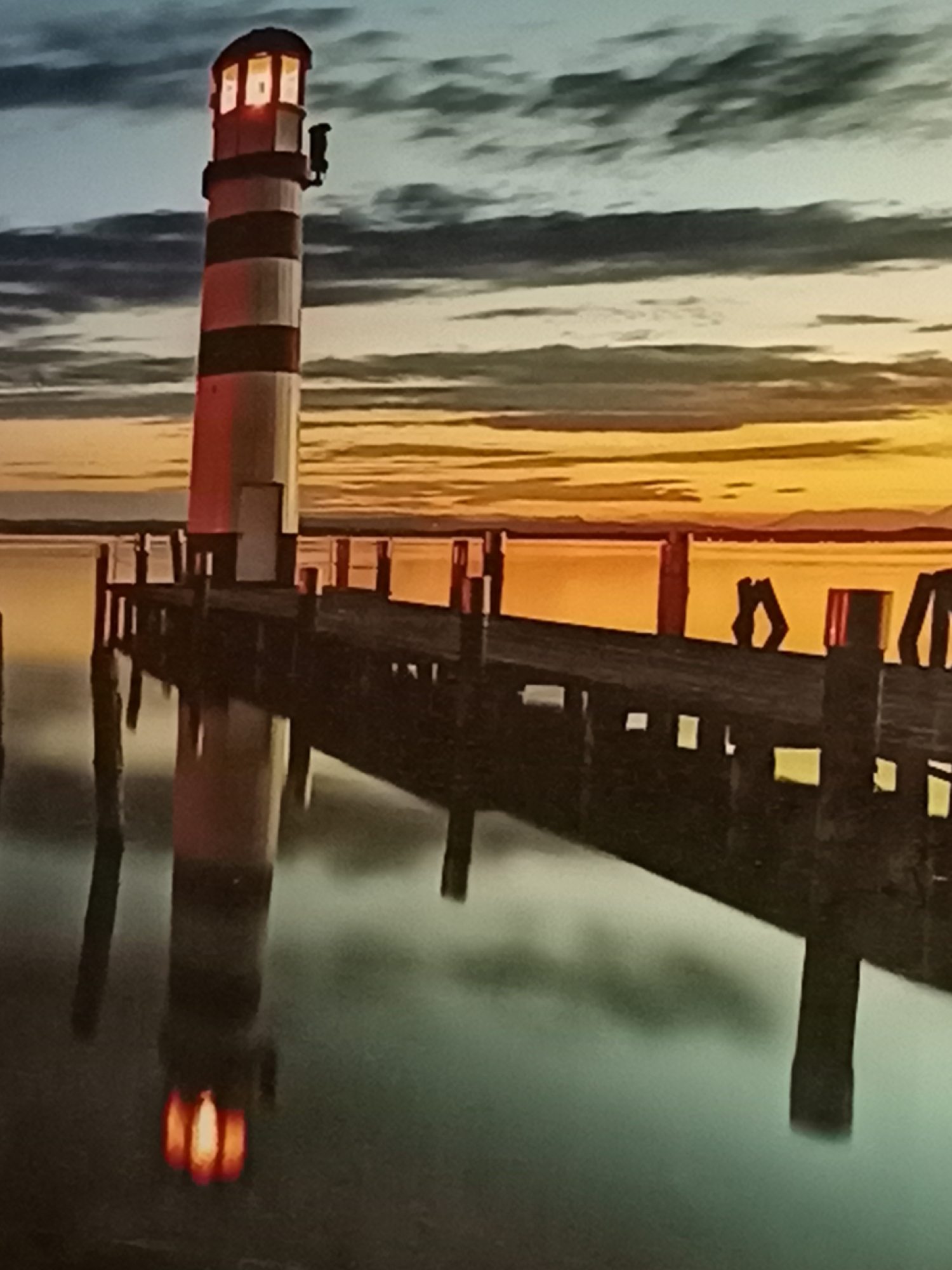 The image shows a striped lighthouse at the end of a pier, reflected in the water below. The sky is a blend of blues and oranges, suggesting either sunrise or sunset. The pier is made of wood and has several posts supporting it. The water is calm and reflects the lighthouse and the sky. The scene is serene and peaceful. There are no people or animals visible in the image. The location is not specified, and there is no indication that it is in Amarillo, United States of America.