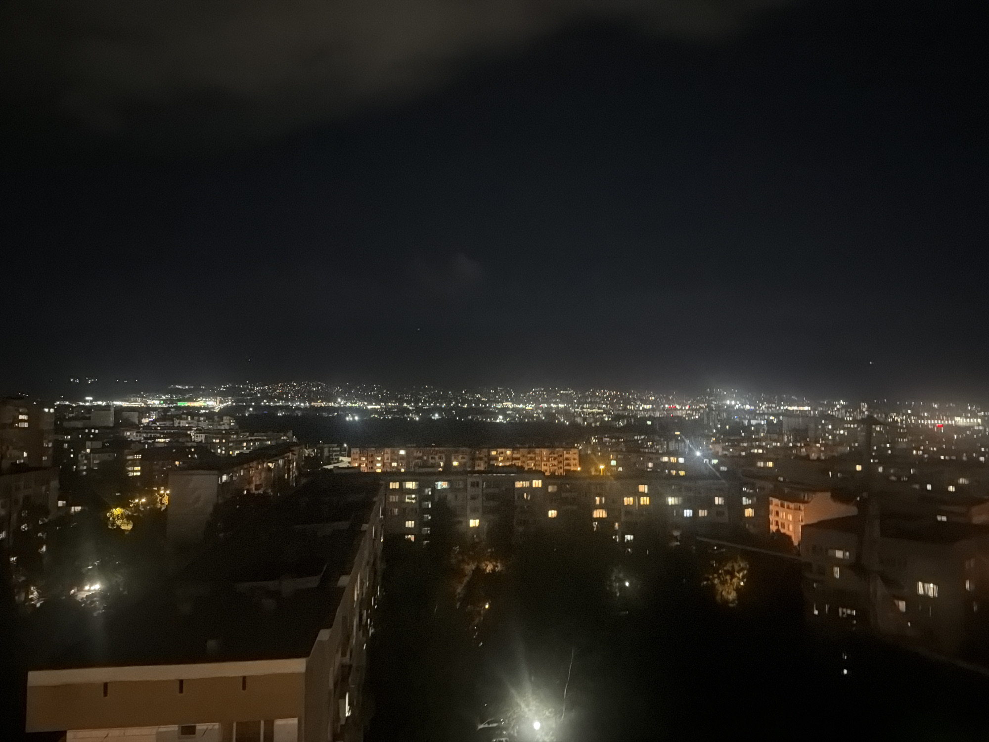 A wide-angle, elevated night view captures the sprawling cityscape of Sofia, Bulgaria. The scene is dominated by a dark night sky with subtle cloud formations overhead. Below, countless artificial lights illuminate a vast urban landscape stretching into the distance. Residential buildings with numerous glowing windows are prominent in the foreground and midground, indicating widespread occupancy. Some closer buildings reveal architectural details such as rooftops and walls, contrasting with the darker areas of unlit foliage and streets between them. Further into the background, the city lights become a dense, shimmering tapestry, outlining the contours of the urban area against the distant horizon. The variety of light colors, primarily white, yellow, and orange, suggests different light sources from homes, streets, and potentially commercial establishments. The sky shows a few extremely faint, ambiguous pinpricks of light. There is no visible human activity or text. The overall impression is one of nocturnal tranquility over a large, active metropolis. The time of day is clearly night, with partial cloud cover.