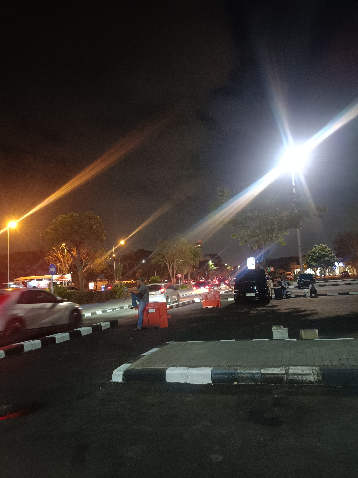 This image depicts a nighttime street scene in Segoro Tambak, Indonesia. The scene is illuminated by bright streetlights that create strong lens flares. Several cars are visible, including a white sedan in the foreground and a black minivan in the mid-ground. A man wearing a grey shirt and blue jeans is seated on an orange traffic barrier, appearing to rest or wait. Another person, also seated, is visible near the black minivan. Trees line the street, and buildings are partially visible in the background. The road surface is asphalt, with painted curbs and marked parking areas. The overall atmosphere is one of a busy urban environment at night.