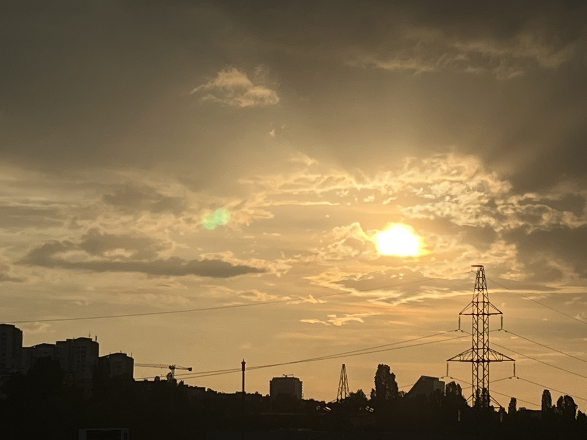 The image captures an urban-suburban landscape in Sofia, Bulgaria, at either sunrise or sunset, characterized by a heavily back-lit scene creating distinct silhouettes. The sky is dominated by a warm, golden-orange glow near the sun, which is obscured by clouds in the upper right quadrant, appearing as a bright, overexposed light source. Away from the sun, the sky transitions to darker grey-brown tones. Stratocumulus clouds are scattered across the sky, with some brightly illuminated from behind, while others appear as dark formations. Two green lens flares are visible: a prominent one in the upper left sky and a fainter one below the sun.
The lower half of the image is completely silhouetted, presenting a dark horizon line composed of structures and vegetation. On the left side of the silhouette, a cluster of multi-story buildings, characteristic of late 20th/early 21st-century Bulgarian urban architecture, is visible. A large construction crane, identifiable by its lattice framework, rises above these buildings. Several horizontal power or communication lines traverse the sky above these structures. Towards the right side, a tall, distinctive electricity transmission pylon is a prominent feature. The pylon is constructed from lattice steelwork, featuring an X-shaped base and upper section, with multiple triangular cross-arms extending outwards. Below and around the pylon, dense clusters of silhouetted trees and other low-rise structures are discernible. Further left from the pylon, another multi-story building is partially visible in silhouette.
The scene depicts a developed area of Sofia, likely a suburban or peripheral district, situated on undulating or slightly hilly terrain, with ongoing construction and visible electrical grid infrastructure. No people are discernible in the image.